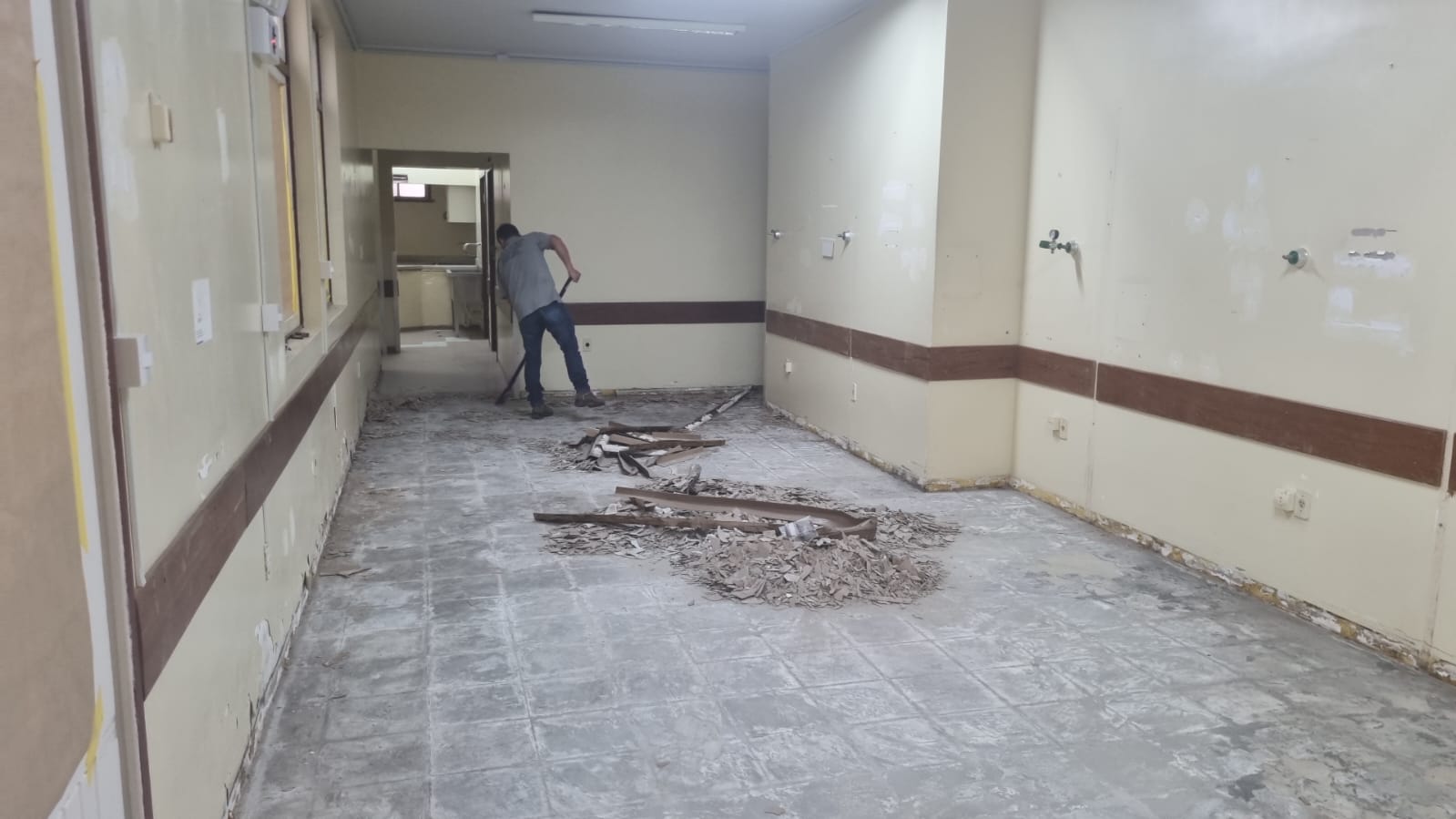 Homem realiza a remoção do piso em uma sala interna de paredes claras com faixas marrons. Há entulhos espalhados pelo chão, como pedaços de revestimento e madeira. O ambiente está visivelmente em reforma, com marcas de equipamentos médicos nas paredes e tomadas expostas, sugerindo que o local pode ter sido um espaço hospitalar ou clínico.
