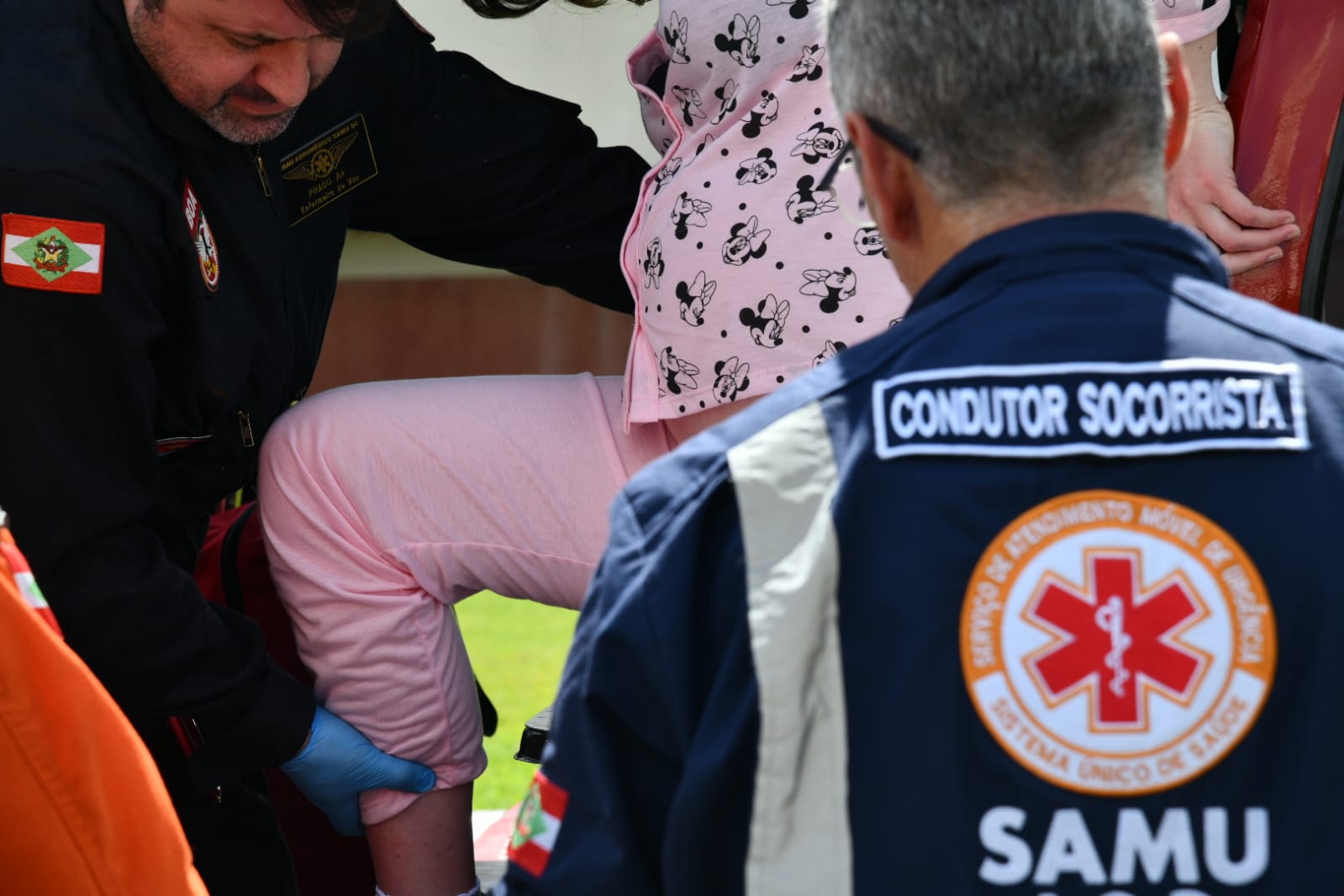 Profissionais do SAMU prestam atendimento a uma gestante vestida com pijama rosa, estampado com personagens. Um socorrista com uniforme azul está de costas, com a inscrição "Condutor Socorrista", enquanto outro profissional segura cuidadosamente a perna da paciente, demonstrando assistência no transporte ou avaliação médica.