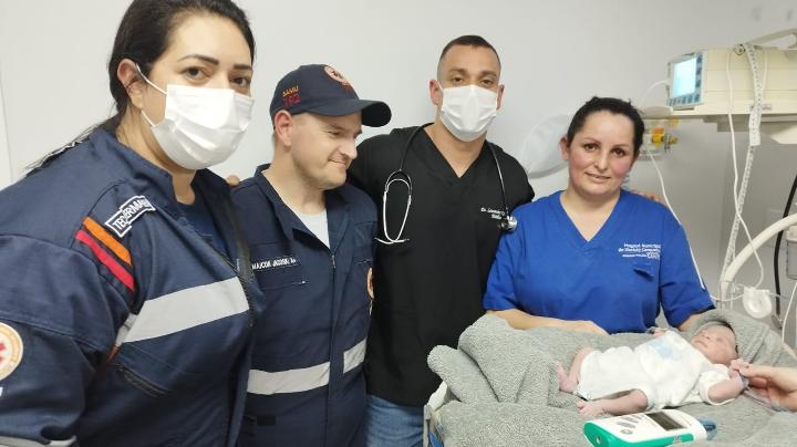 Profissionais de saúde e socorristas do SAMU ao lado de recém-nascido enrolado em manta sobre maca hospitalar, em ambiente de atendimento neonatal.
