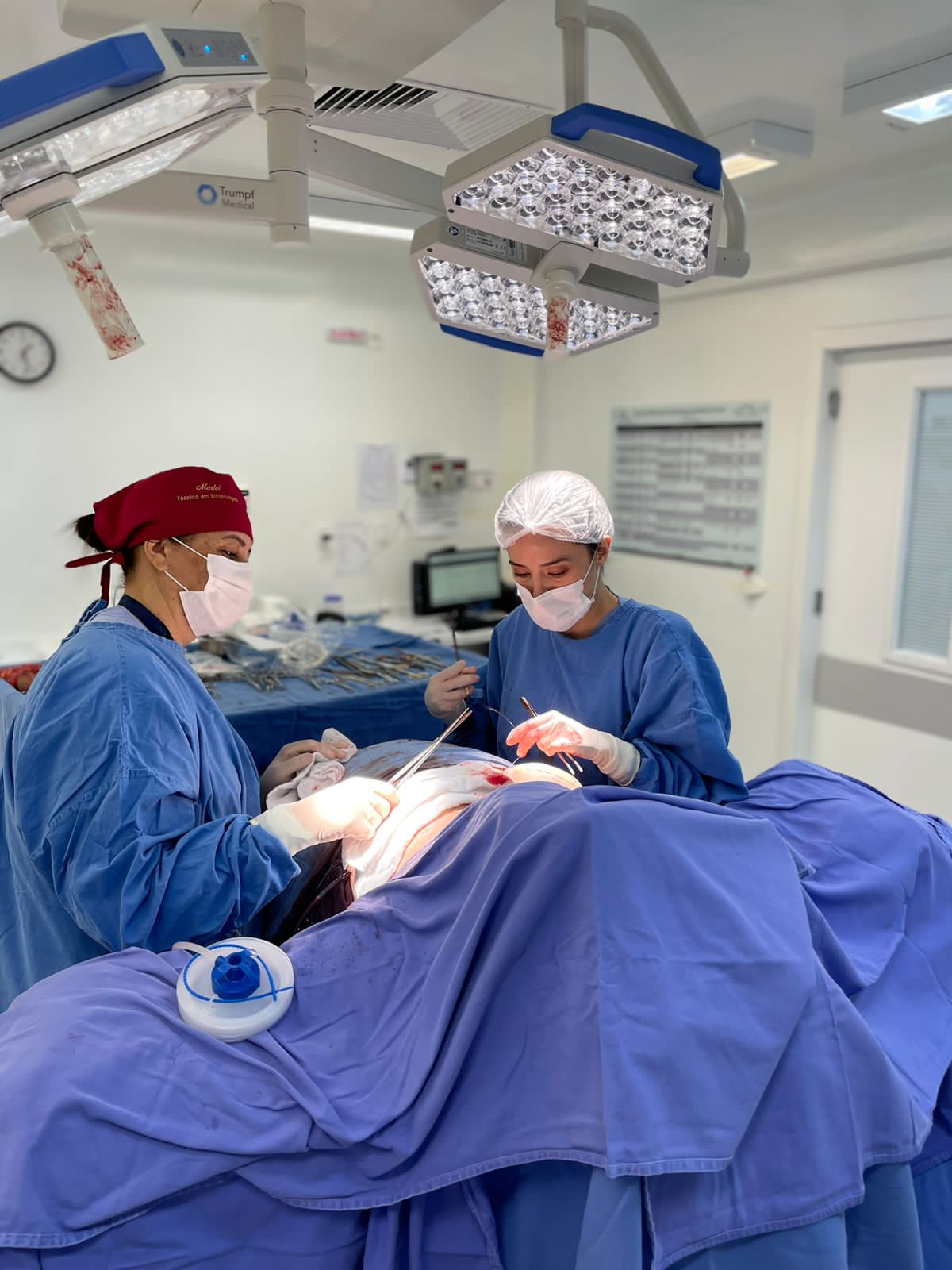 Duas profissionais de saúde, vestindo roupas cirúrgicas azuis, toucas e máscaras, realizam procedimento em paciente coberto por campos estéreis, sob iluminação de focos cirúrgicos no centro cirúrgico.