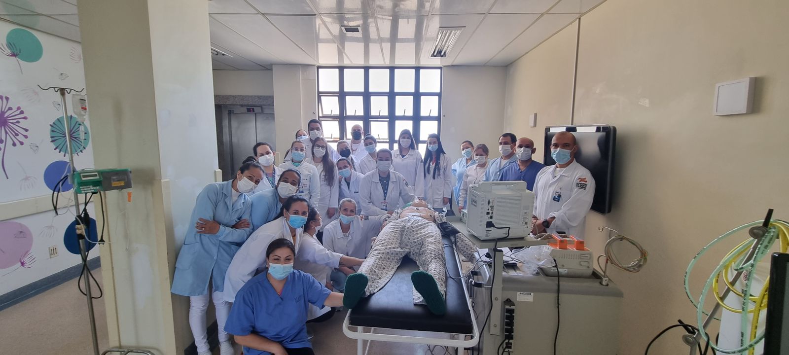A imagem mostra um grupo numeroso de profissionais de saúde, todos usando máscaras e jalecos ou uniformes hospitalares, reunidos em uma sala de atendimento médico. No centro, há uma maca com um paciente deitado (provavelmente um manequim de simulação médica, dado o contexto e a disposição), rodeado por equipamentos hospitalares como monitor multiparamétrico, cabos e tubos.  A iluminação é natural, vinda de uma grande janela ao fundo. À esquerda, parte da parede tem uma decoração com desenhos coloridos, sugerindo que o ambiente pode estar em uma ala pediátrica ou humanizada. À direita, há equipamentos médicos fixados e móveis, além de um monitor de parede.  O clima geral parece ser de treinamento, capacitação ou registro de equipe, mais do que de atendimento emergencial.