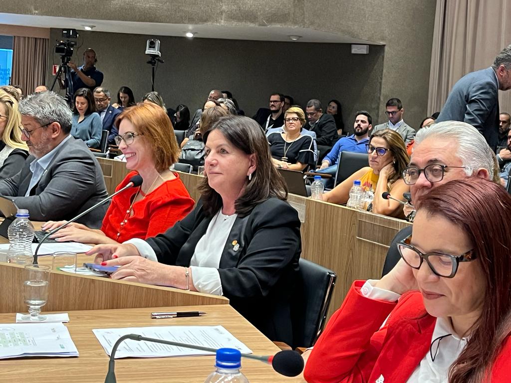 A imagem mostra um ambiente de reunião formal, possivelmente uma audiência pública, sessão legislativa ou conferência.  Em primeiro plano, há várias pessoas sentadas atrás de mesas de madeira clara, com microfones e documentos à frente. Algumas usam roupas sociais em tons de vermelho, preto e branco, e a maioria está concentrada, ouvindo ou interagindo. No canto inferior esquerdo, vê-se uma garrafa de água e um copo vazio.  Ao fundo, mais participantes e observadores estão sentados, alguns com laptops abertos. Também é possível notar câmeras de gravação e um cinegrafista, o que indica que o evento está sendo registrado. O clima geral é sério e profissional, mas com momentos de interação mais descontraída entre os presentes.