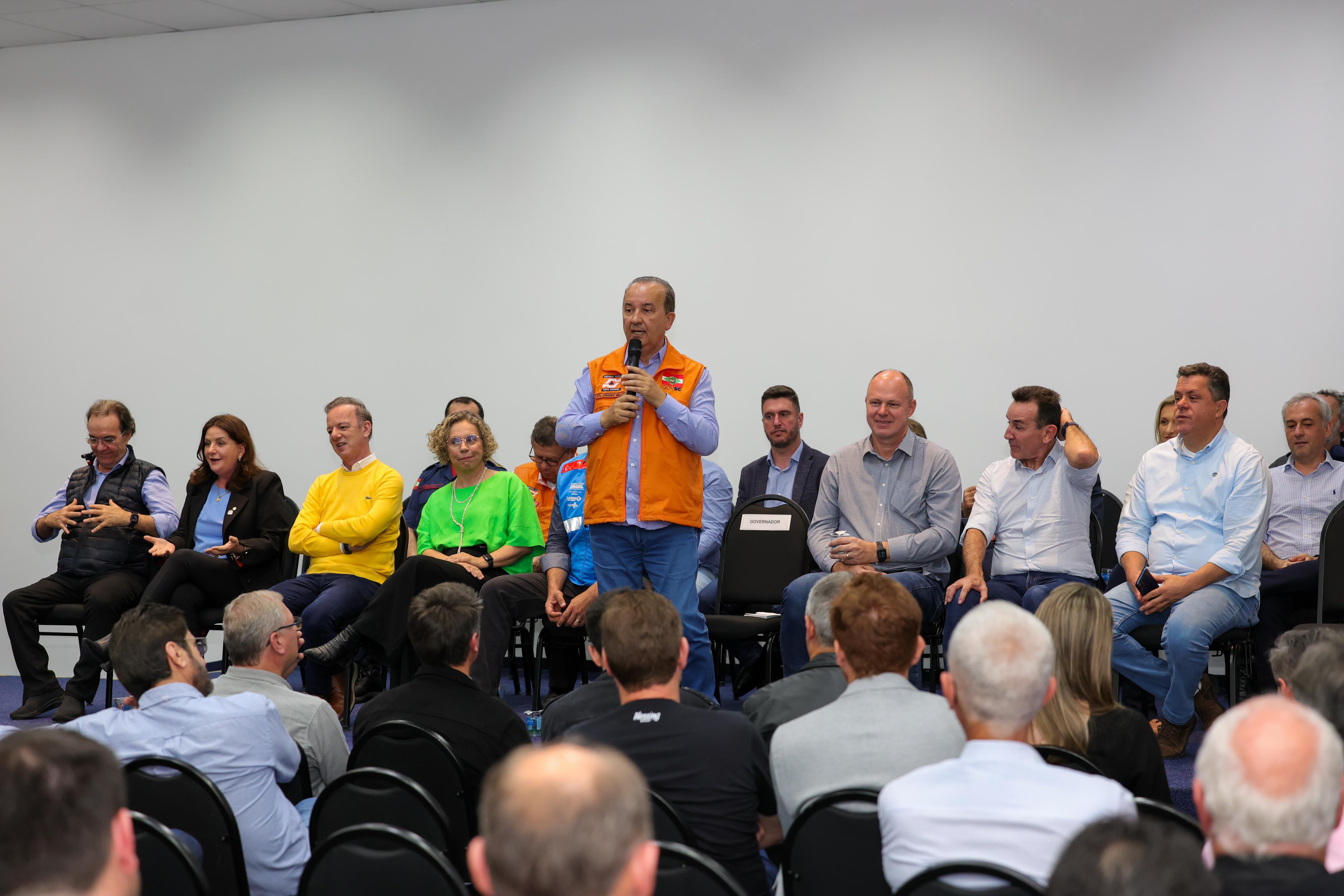 Homem de colete laranja da defesa civil falando ao microfone para plateia, com diversas autoridades sentadas atrás dele durante reunião no Vale do Itajaí.