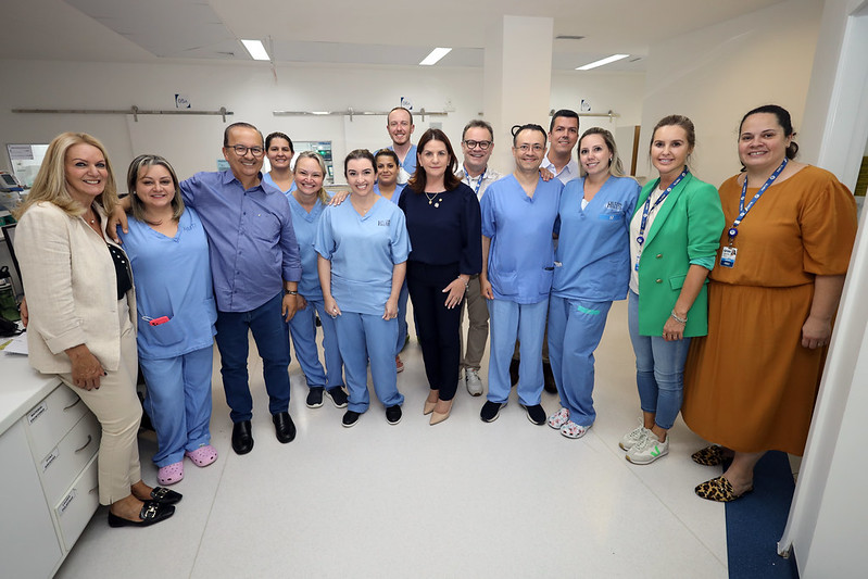 Grupo de profissionais da saúde e representantes em ambiente hospitalar. Eles posam sorrindo para a foto em um corredor claro e moderno. A maioria veste uniforme azul hospitalar, enquanto alguns usam roupas sociais e crachás.