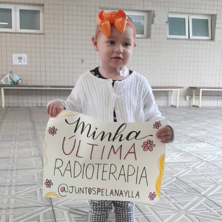 Criança pequena, sem cabelos e usando laço laranja na cabeça, veste suéter branco e segura um cartaz com a frase “Minha última radioterapia @juntospelanaylla”, decorado com flores e desenhos coloridos. A cena transmite alegria e superação, e ocorre em um espaço interno com paredes de azulejo claro e bancos ao fundo.