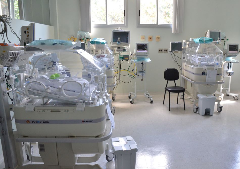 Sala de UTI Neonatal equipada com várias incubadoras para recém-nascidos, todas conectadas a monitores e aparelhos de suporte à vida. A sala é bem iluminada, com paredes claras e janelas ao fundo. Cadeiras e equipamentos adicionais complementam o ambiente clínico e organizado.