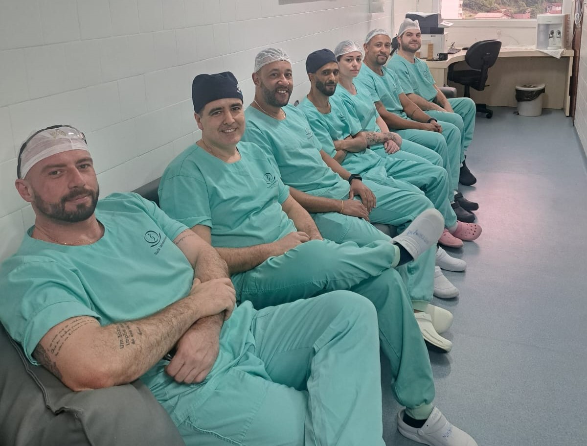 Equipe médica de ortopedia sentada em fila, usando uniformes verdes e toucas cirúrgicas.