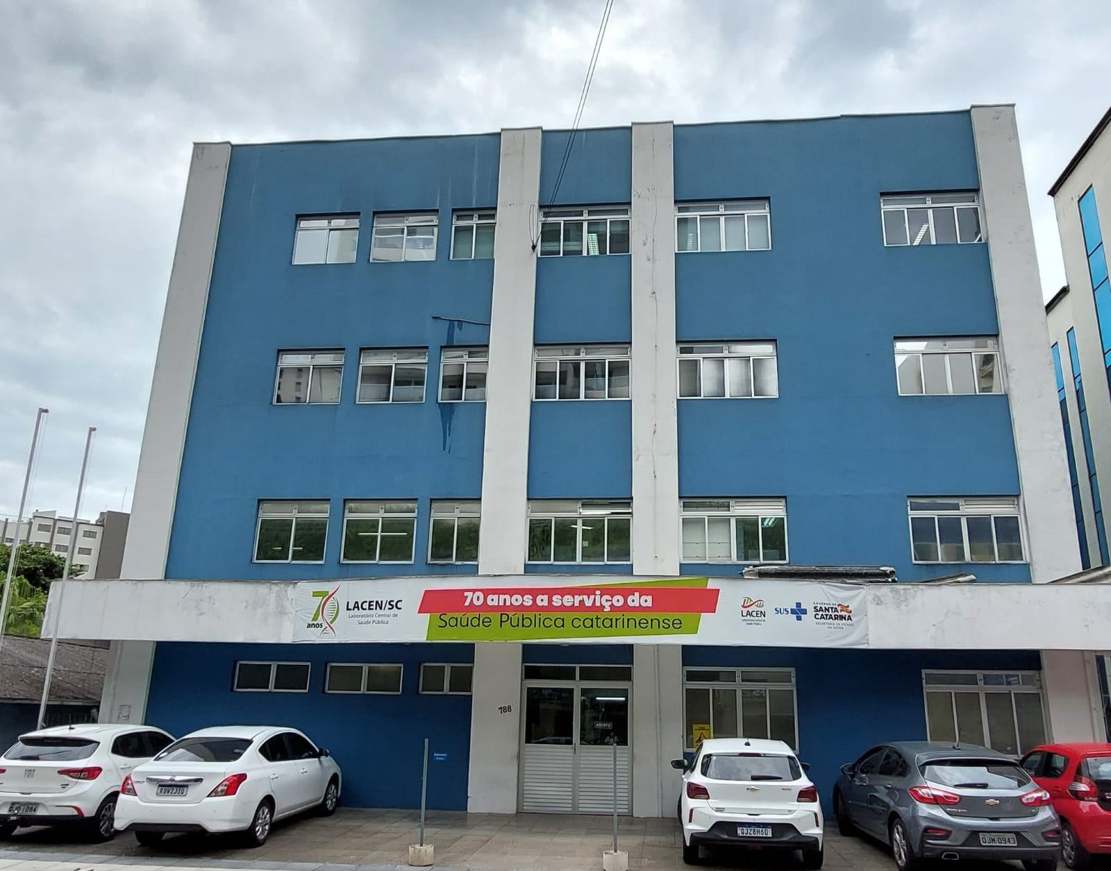 Prédio de três andares com fachada azul e detalhes verticais brancos, sede do LACEN/SC (Laboratório Central de Saúde Pública de Santa Catarina). Há uma faixa na entrada destacando “70 anos a serviço da Saúde Pública catarinense” com logotipos do LACEN, SUS e Governo de Santa Catarina. Na frente do prédio, há cinco carros estacionados. O céu está nublado, conferindo iluminação difusa à cena.