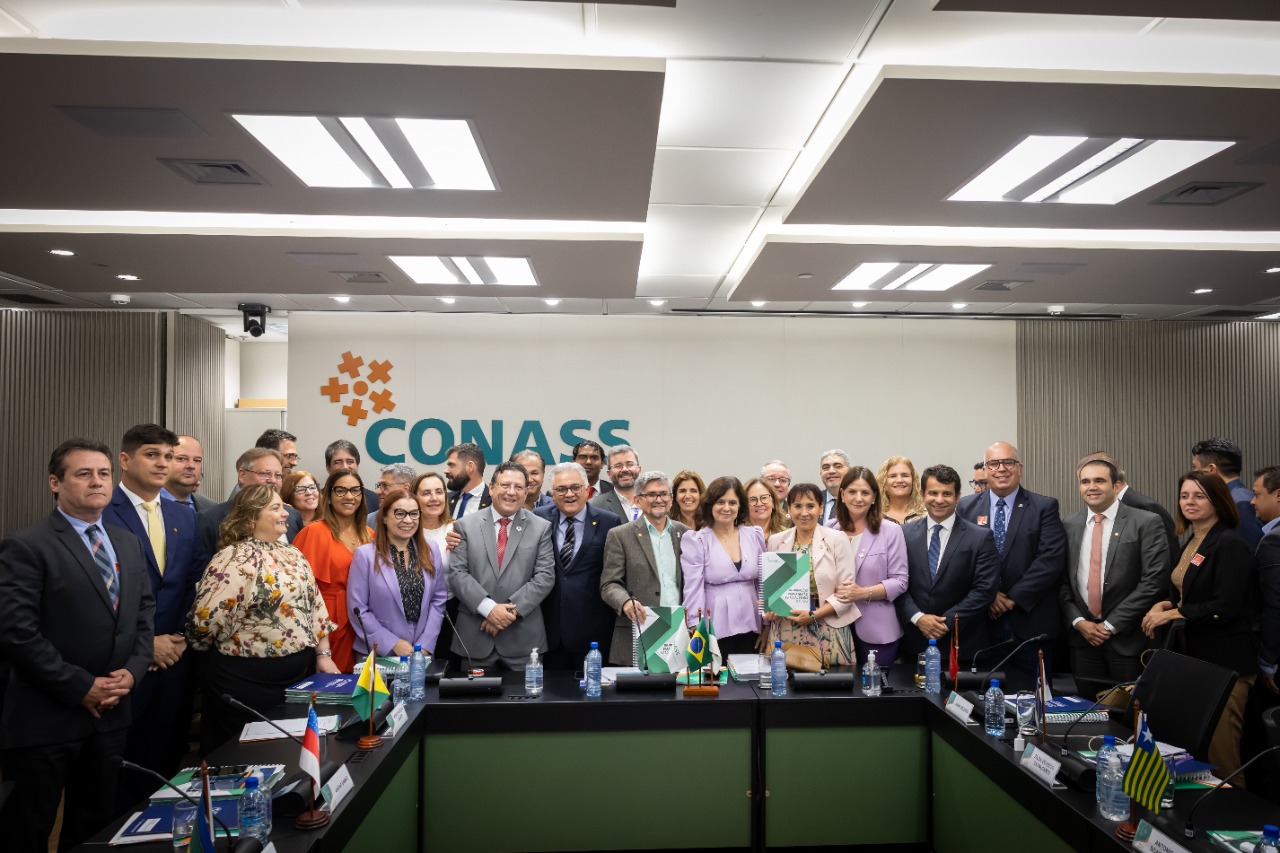 : Foto de um grande grupo de pessoas posando para uma foto oficial em uma sala de reuniões. Ao fundo, há a logomarca do CONASS (Conselho Nacional de Secretários de Saúde) em uma parede branca. A maioria dos participantes está de terno, blazer ou trajes formais, com expressões sorridentes. Algumas pessoas seguram pastas com o brasão do Brasil. Sobre a mesa à frente, há garrafas de água, crachás e pequenas bandeiras estaduais. A iluminação é moderna e o ambiente sugere um evento institucional ou reunião de alto nível do setor público de saúde.