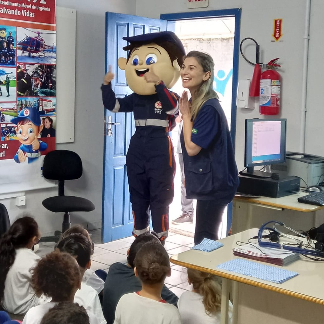 Profissional do SAMU apresenta mascote em tamanho real para um grupo de crianças em uma sala de aula. A figura do mascote, sorridente e uniformizada, faz gestos com as mãos, enquanto a profissional ao lado sorri entusiasmada. As crianças observam sentadas no chão, atentas à apresentação educativa sobre serviços de emergência.