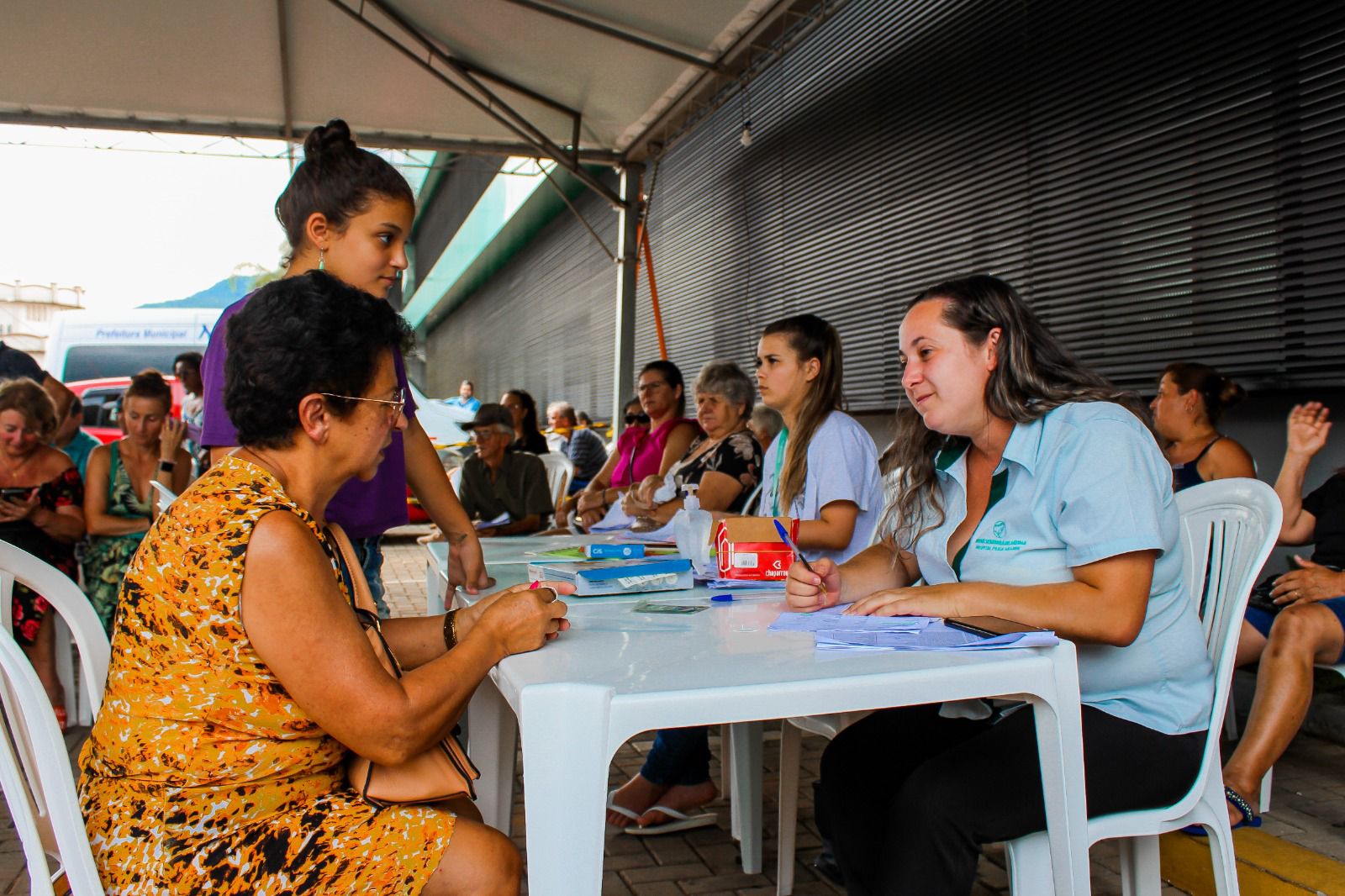 Atendimento comunitário em ambiente externo e coberto, com profissionais da saúde conversando com moradores sentados em cadeiras plásticas. Uma mulher com camisa institucional registra informações enquanto outra mulher, de vestido amarelo, é atendida. Ao fundo, outras pessoas aguardam ou também estão em atendimento.