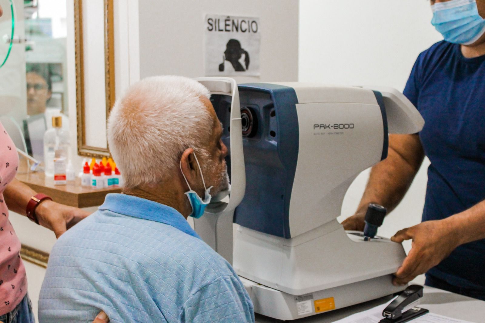 Idoso realiza exame oftalmológico com equipamento automatizado (autorefrator) operado por profissional da saúde. O exame acontece em ambiente clínico, com sinalização de “Silêncio” na parede ao fundo. O paciente está com a máscara abaixada e veste camisa azul clara.