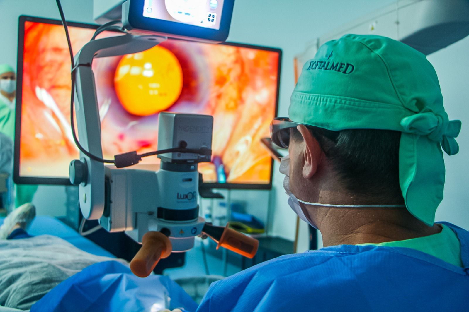 Médico realiza cirurgia oftalmológica assistida por microscópio cirúrgico e monitor de alta definição, onde é exibida em detalhe a imagem ampliada do olho do paciente. O profissional veste touca verde com a inscrição "HOSPITALMED" e avental cirúrgico azul, em ambiente estéril de centro cirúrgico.