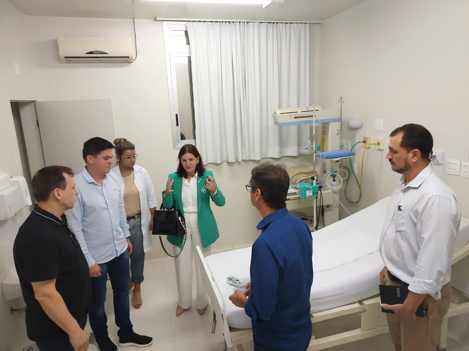 Grupo de pessoas visita um quarto hospitalar com leito clínico. Entre os presentes está uma mulher de blazer verde e calça branca, falando com os demais. A sala é equipada com cama hospitalar, equipamentos médicos e cortinas brancas.