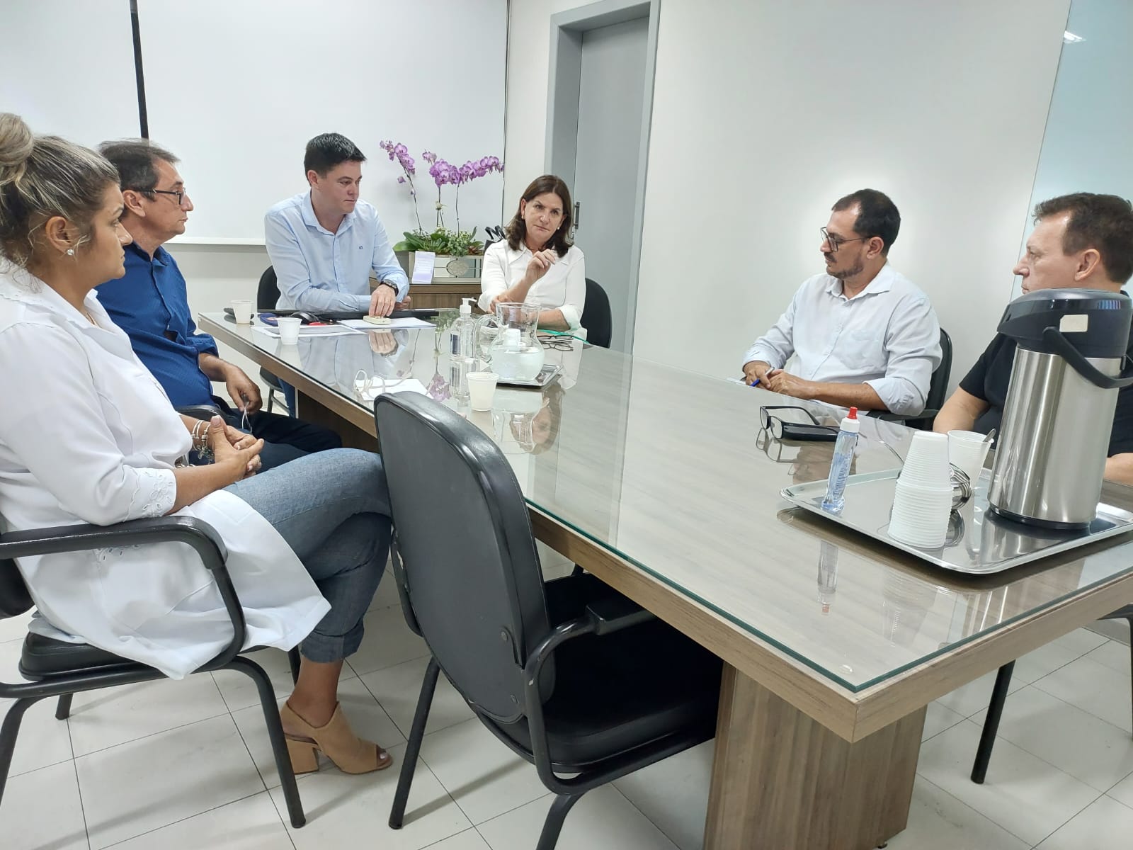 Grupo de sete pessoas participa de reunião ao redor de uma mesa de vidro em uma sala clara e moderna. Entre eles, há profissionais de saúde e gestores, com documentos e materiais de apoio sobre a mesa. Ao fundo, um vaso com orquídeas decora o ambiente.