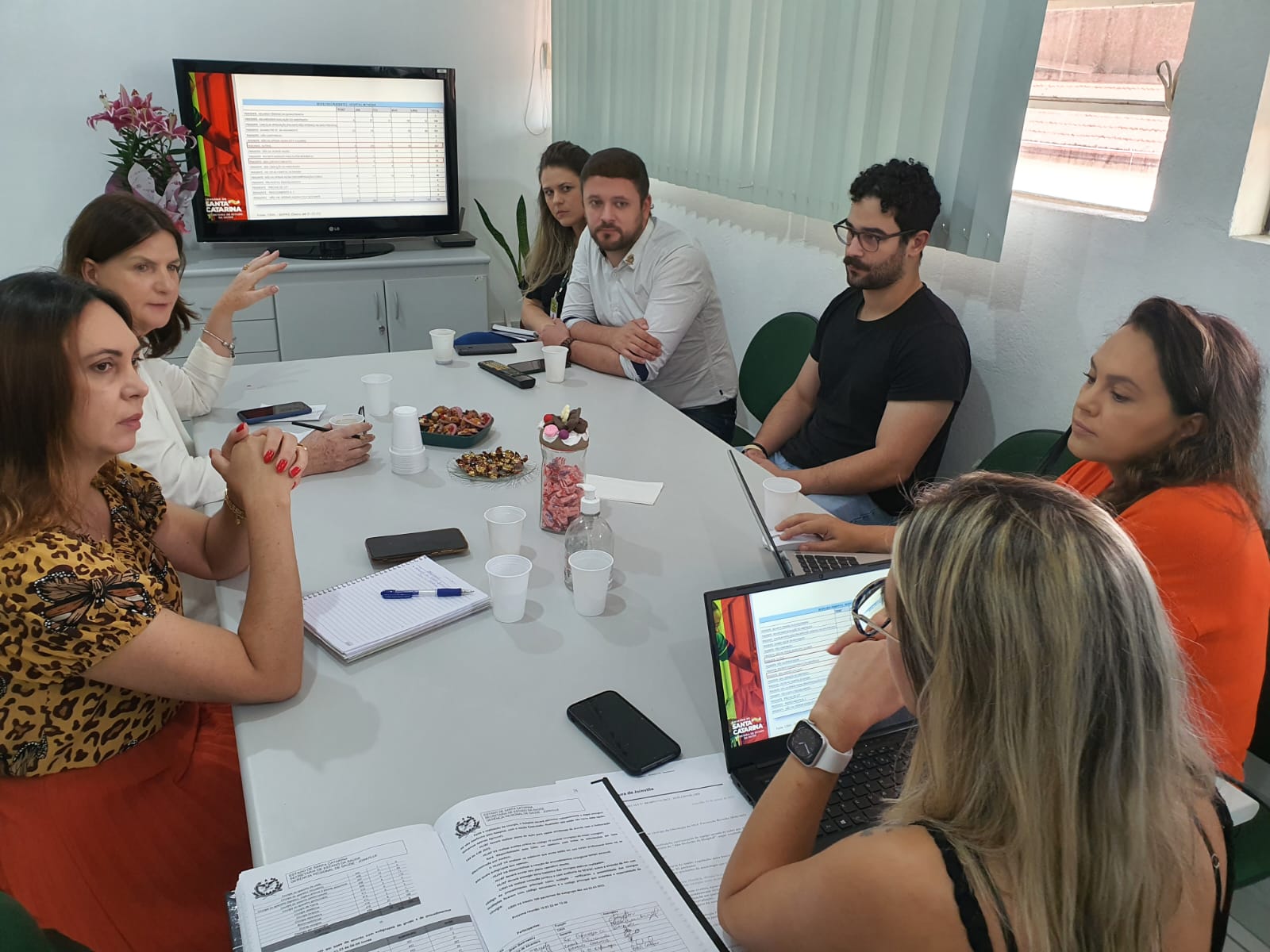 Reunião de equipe com oito pessoas sentadas ao redor de uma mesa retangular discutindo documentos e planilhas exibidas em notebooks e em uma TV ao fundo, que mostra uma tabela com o logotipo do Governo de Santa Catarina. Há copos, cadernos, celulares e pratos com doces sobre a mesa.