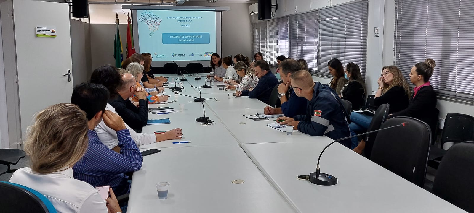 Sala de reunião com cerca de 20 pessoas sentadas ao redor de uma longa mesa branca, participando de um encontro formal. No fundo, há uma tela projetando a apresentação do "Projeto de Fortalecimento da Gestão Estadual do SUS", com logotipos de instituições e a identificação da Secretaria de Estado da Saúde de Santa Catarina. Alguns participantes fazem anotações enquanto ouvem atentamente.