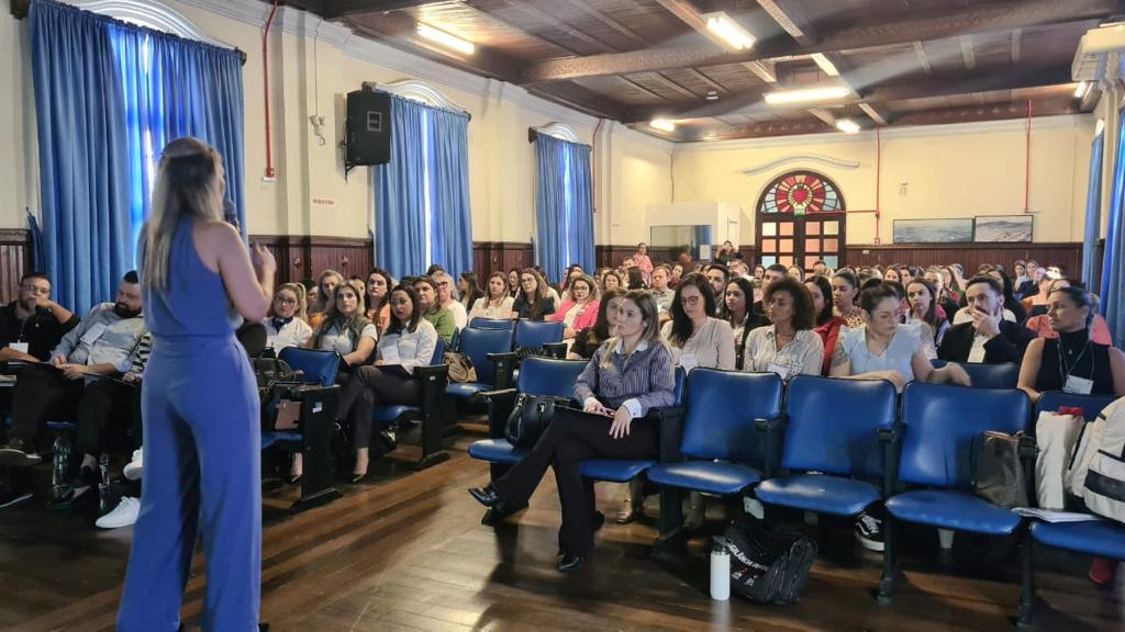 Auditório com várias pessoas sentadas prestando atenção à palestrante em pé, de costas para a câmera. O local tem cadeiras azuis, cortinas azuis e teto de madeira, com boa iluminação.