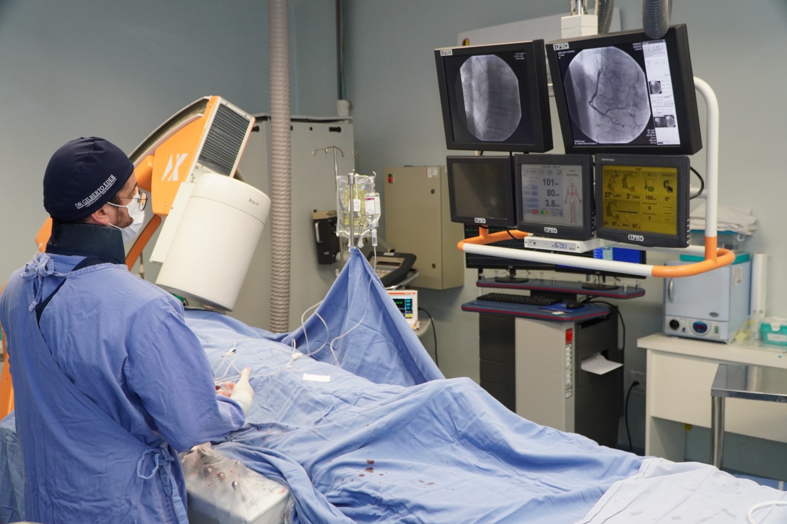 Profissional de saúde realizando procedimento de hemodinâmica, com paciente coberto por campos cirúrgicos azuis e monitores exibindo imagens e dados cardíacos.