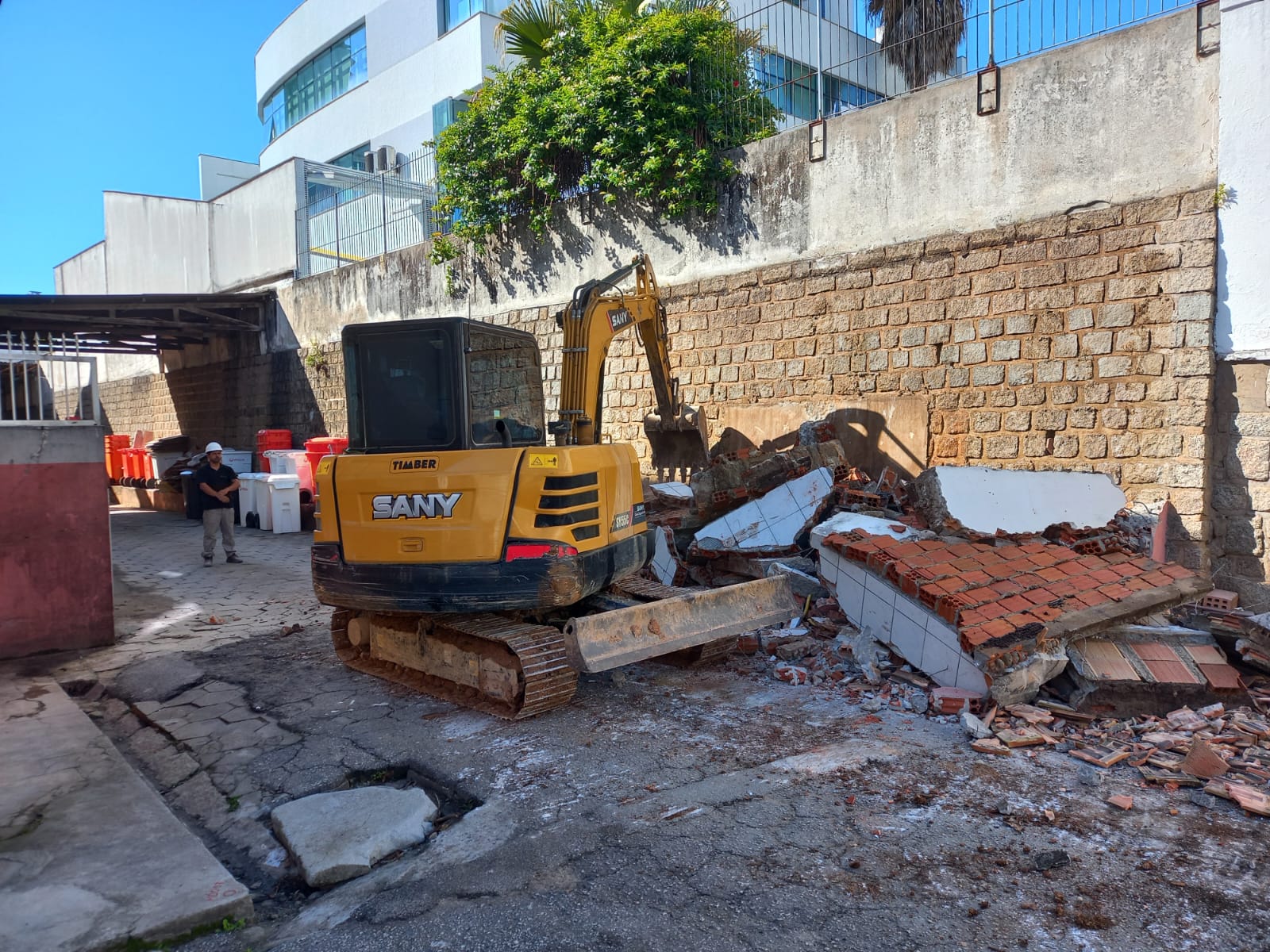 Uma escavadeira amarela da marca SANY está em operação demolindo uma estrutura de alvenaria com tijolos e azulejos, encostada a um muro de pedras. Ao lado, um trabalhador com capacete branco observa a cena. Há contêineres brancos e laranjas organizados sob uma cobertura à esquerda, e ao fundo, vê-se um prédio moderno de vidro. A imagem mostra uma etapa de obras ou reestruturação urbana.