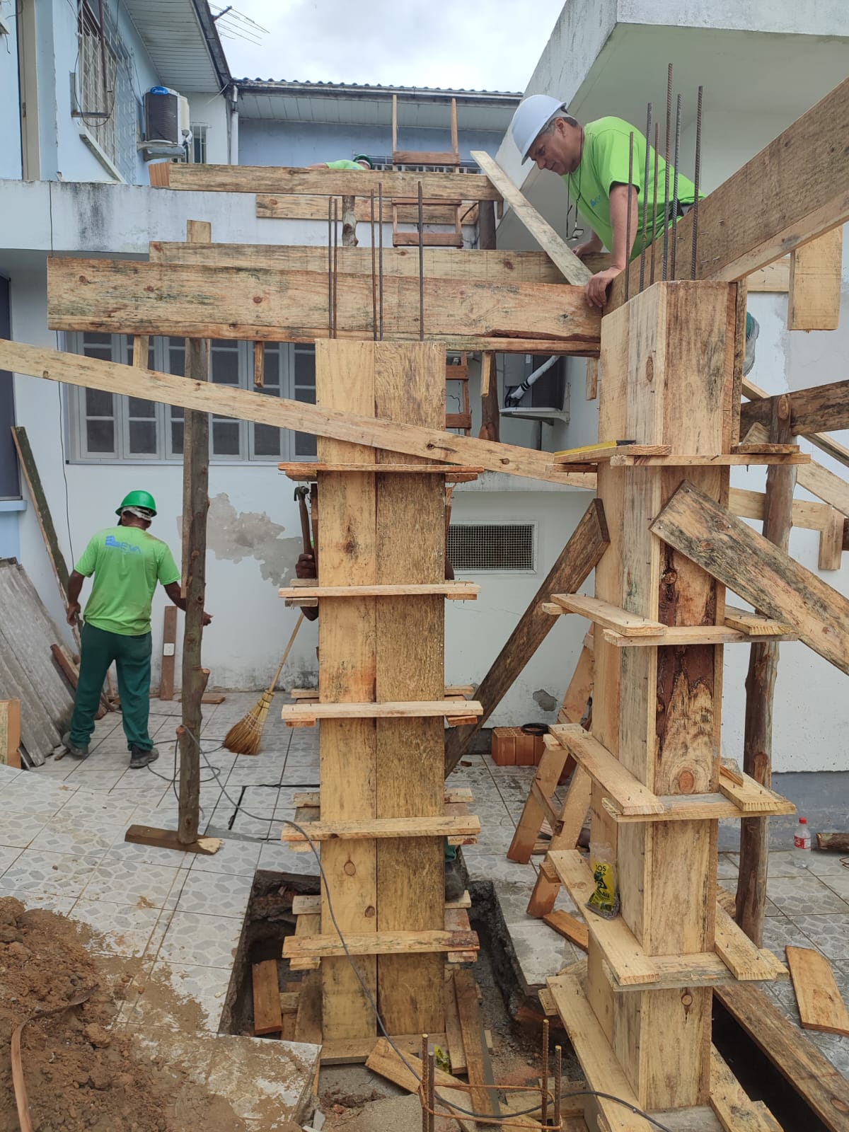 Três trabalhadores com uniformes verdes e capacetes atuam na montagem da estrutura de madeira para colunas de concreto em uma obra. Um deles está no nível superior, apoiado na estrutura, enquanto os outros dois estão no térreo. A imagem mostra o processo de escoramento e preparação para concretagem em um prédio em construção.