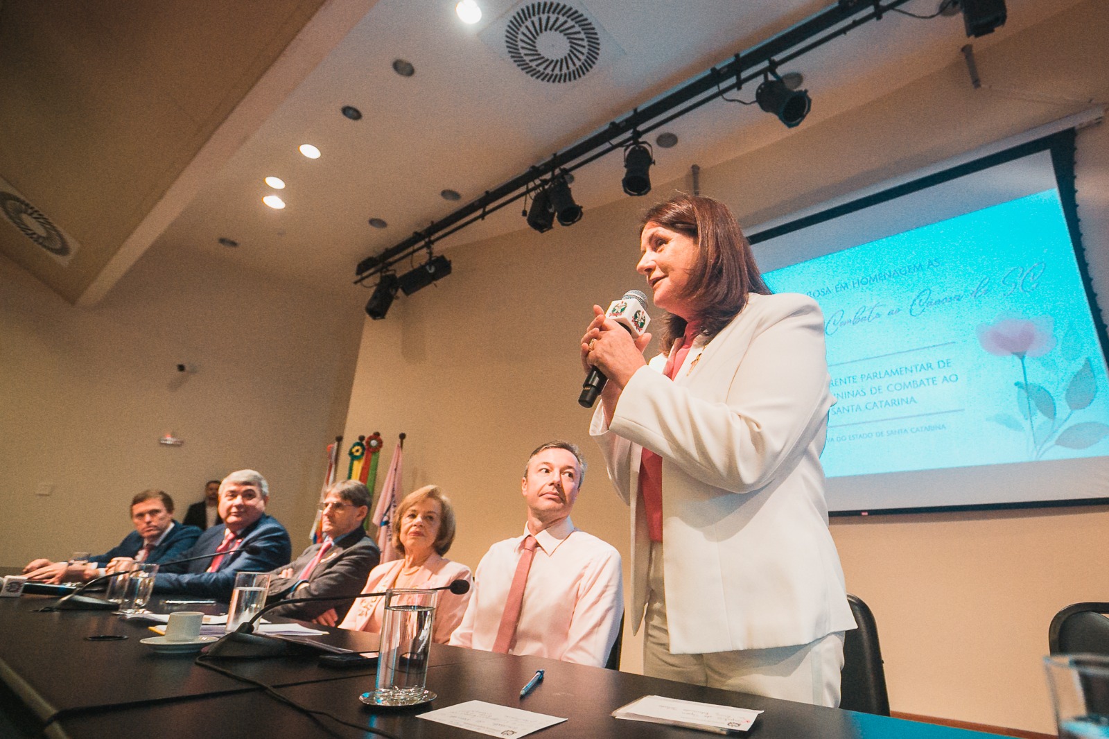 Mulher em pé, vestindo terno branco, falando ao microfone durante evento de conscientização sobre câncer, com painel de convidados sentados e tela projetada ao fundo.