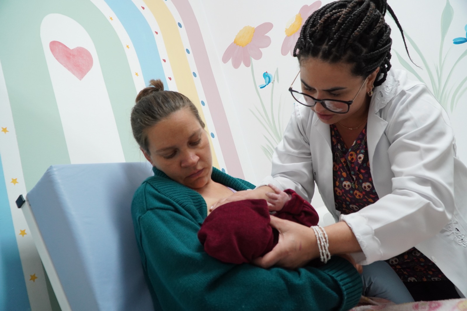 Profissional de saúde auxilia mãe no posicionamento de recém-nascido para amamentação em ambiente colorido com pintura de arco-íris e flores na parede.