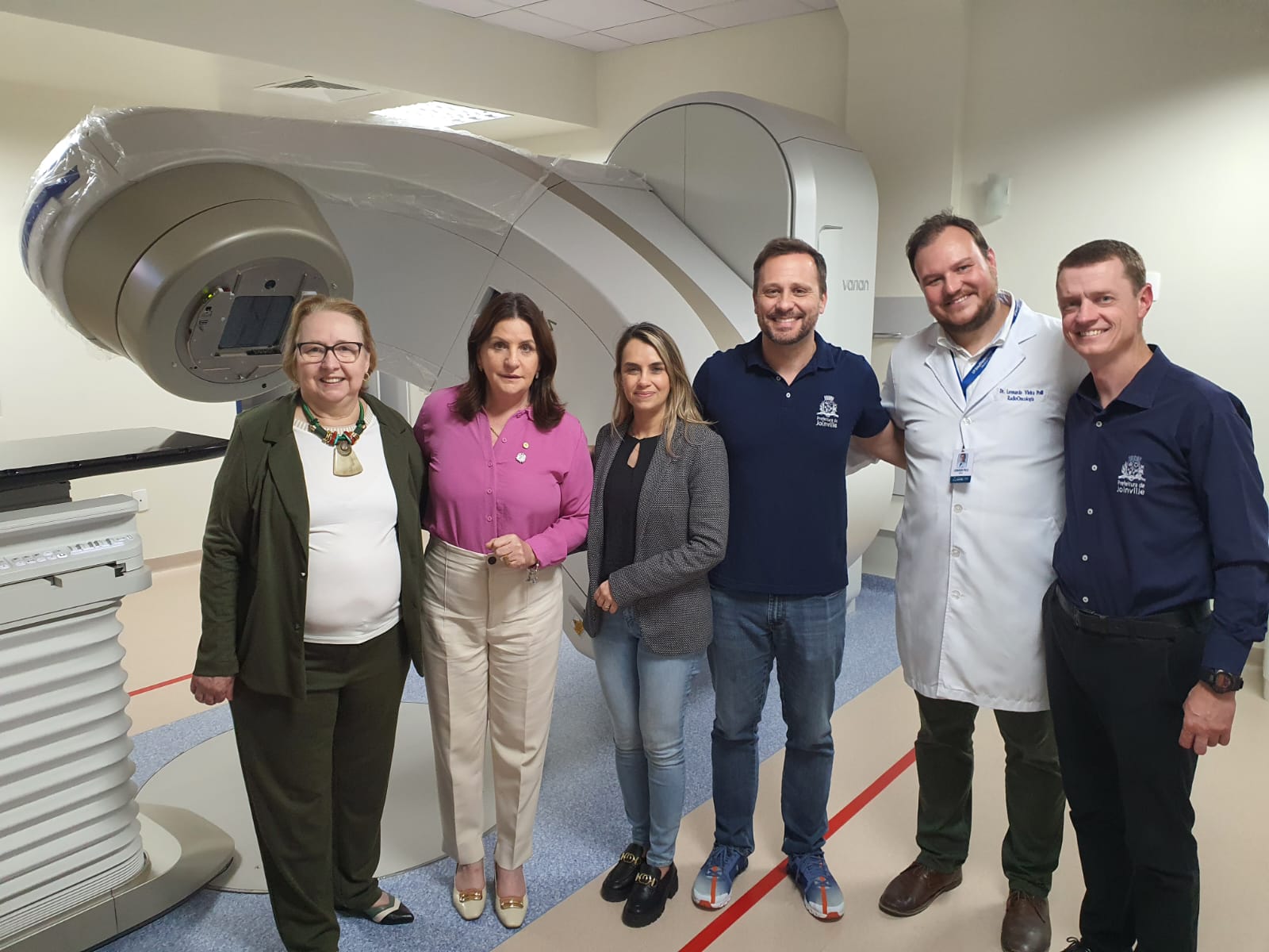 Grupo de seis pessoas posa sorridente em frente a um moderno equipamento de radioterapia em uma sala clínica. O aparelho é grande e ocupa boa parte do fundo da imagem. Entre os presentes, há profissionais da saúde e representantes administrativos, alguns usando jaleco branco ou camisa com logotipo institucional.