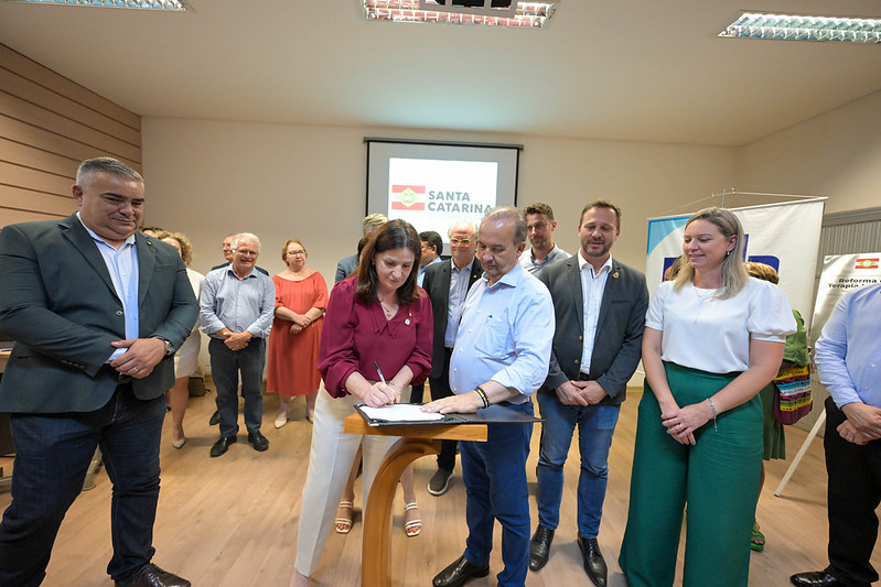 Mulher assinando documento em um púlpito de madeira enquanto é acompanhada por diversas autoridades e servidores públicos. Ao fundo, há uma projeção com o logotipo do Governo de Santa Catarina e pessoas observando o ato. Todos estão em um ambiente interno com iluminação fluorescente e piso de madeira.