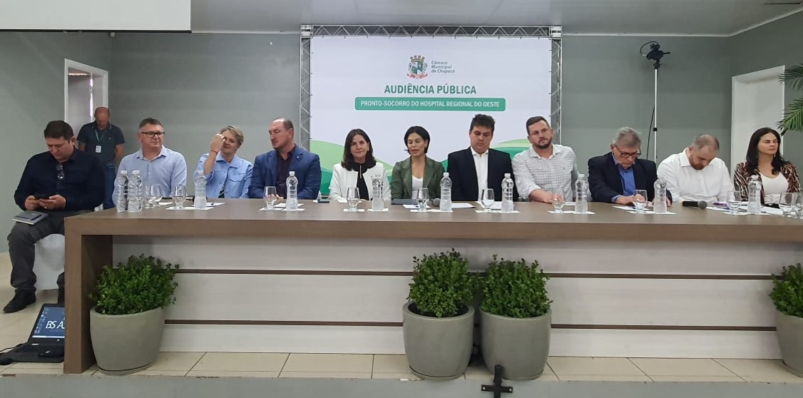Onze pessoas sentadas lado a lado em uma mesa de autoridades durante audiência pública. Atrás, um painel da Câmara Municipal de Chapecó com os dizeres “Audiência Pública – Pronto-Socorro do Hospital Regional do Oeste”. Todos usam roupas formais; à frente, copos e garrafas d'água estão dispostos sobre a mesa, e vasos com plantas ornamentam a frente do palco.