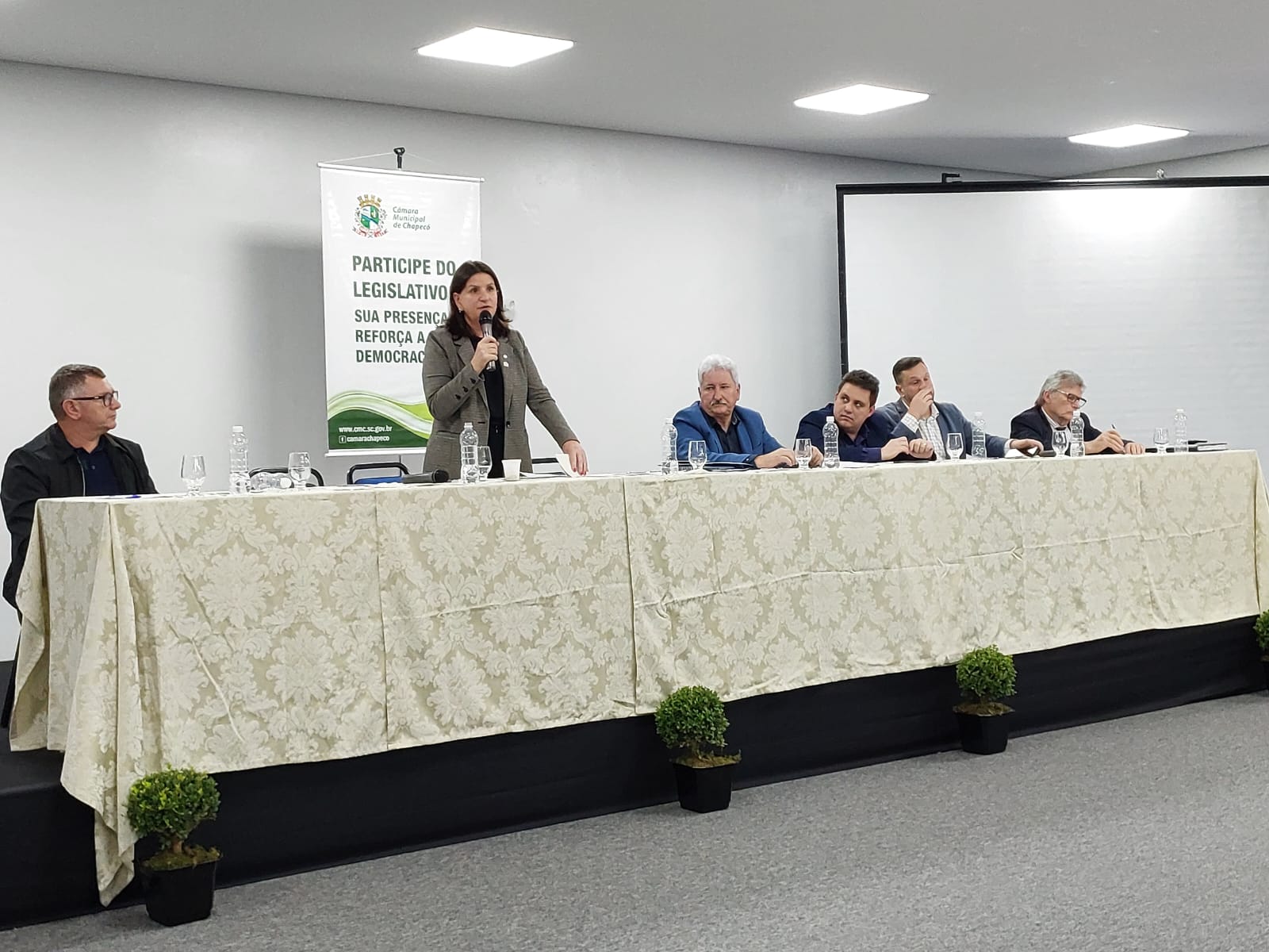 Autoridades e representantes participam de audiência pública sobre o Hospital da Criança de Chapecó. Seis pessoas estão sentadas à mesa principal, enquanto uma mulher fala ao microfone em pé. Ao fundo, banner da Câmara de Chapecó com os dizeres ‘Participe do Legislativo. Sua presença reforça a democracia.
