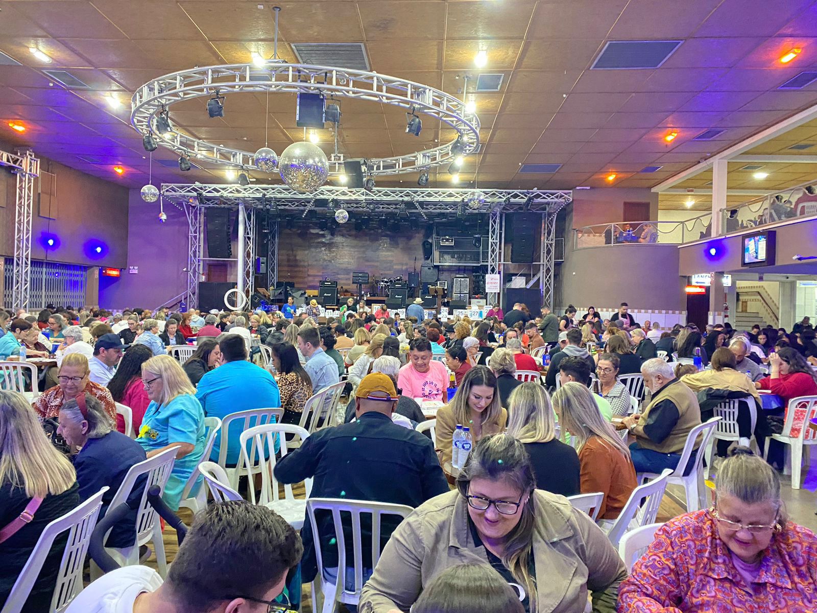 Salão amplo lotado de pessoas participando de um bingo beneficente. As pessoas estão sentadas em cadeiras brancas ao redor de mesas, concentradas em suas cartelas. No fundo, há um palco com equipamentos de som e iluminação.
