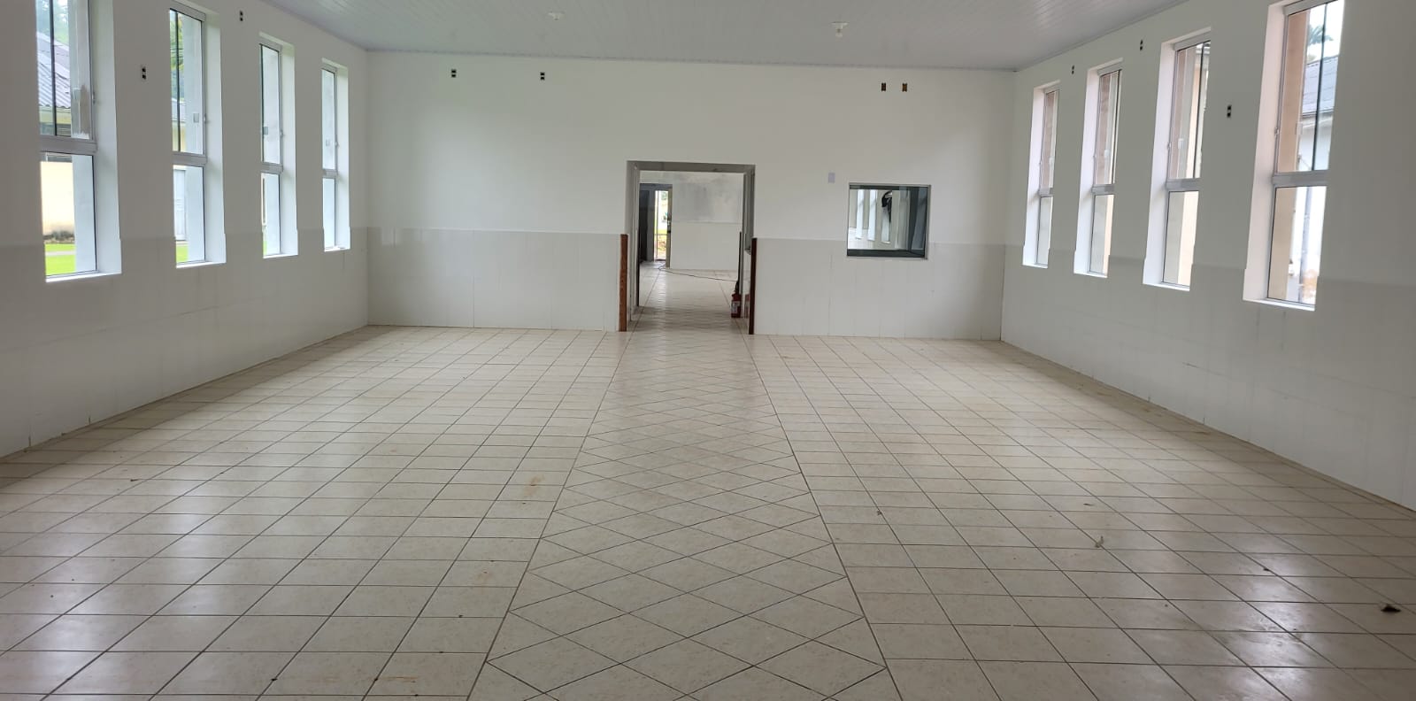 Sala ampla e vazia com piso de cerâmica clara disposta em padrão diagonal, paredes brancas revestidas até a metade com azulejos brancos e teto de forro em PVC branco. Nas laterais, fileiras de janelas altas e retangulares com esquadrias brancas permitem entrada abundante de luz natural. Ao fundo, há uma porta larga que dá acesso a outro corredor e uma janela interna quadrada com moldura metálica, sugerindo um balcão de atendimento.