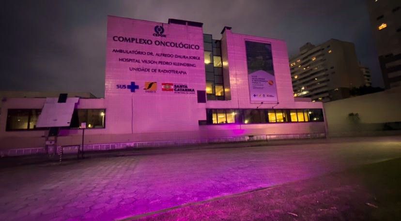 Fachada do Complexo Oncológico Dr. Alfredo da Silva Jorge (CEONC) iluminada com luz rosa à noite, em apoio à campanha Outubro Rosa. O prédio exibe logotipos do SUS, Governo de Santa Catarina e Hospital Universitário. Ao fundo, há edifícios residenciais sob o céu escuro.