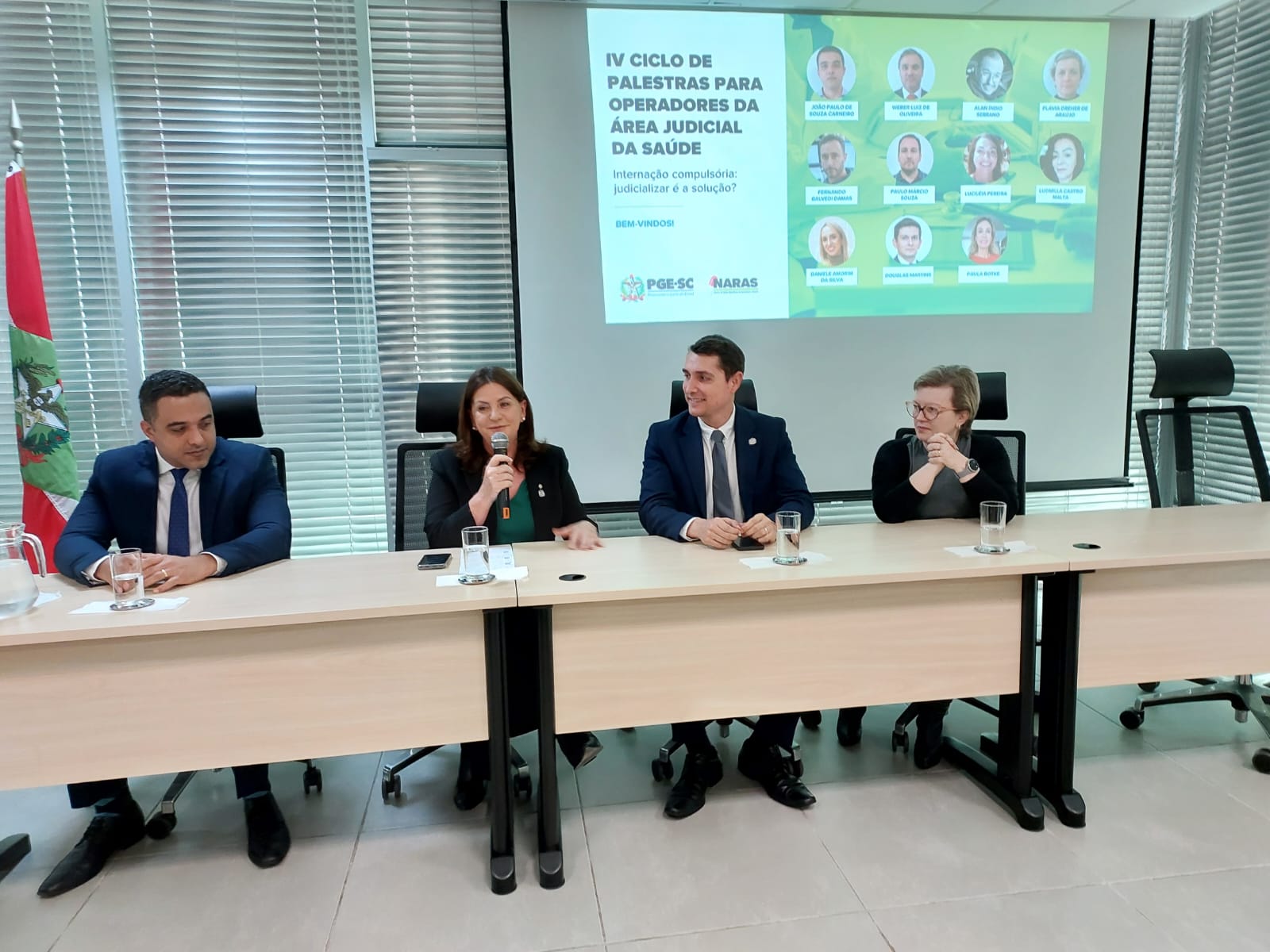 Quatro pessoas sentadas atrás de uma mesa, em auditório, participando do IV Ciclo de Palestras para Operadores da Área Judicial da Saúde, com telão ao fundo exibindo informações do evento e fotos dos palestrantes.