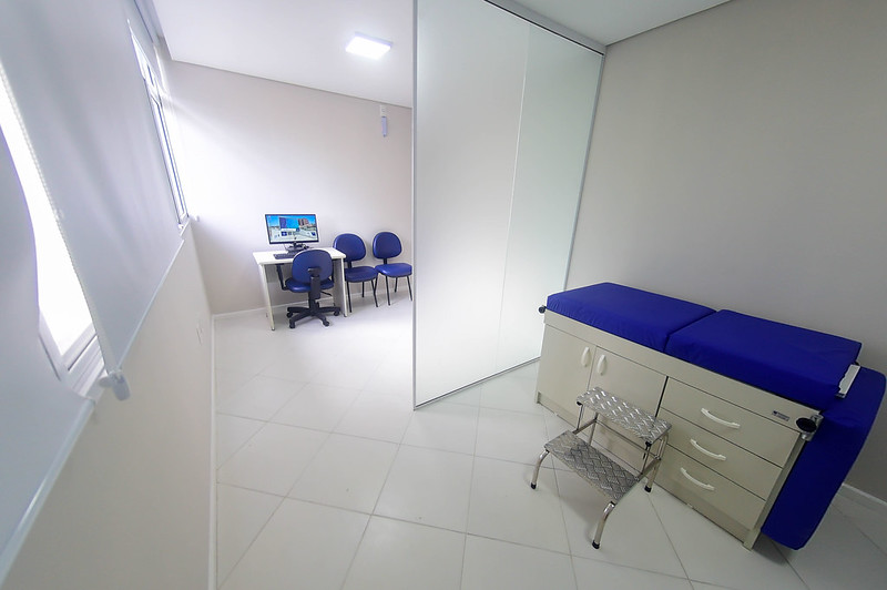 Consultório médico infantil com decoração simples e ambiente limpo. À direita, há uma maca com estofado azul, ao lado de um escadote metálico. Do outro lado da divisória, vê-se uma mesa com computador, três cadeiras azuis e uma cadeira de rodinhas, posicionadas próximo à janela com persianas fechadas. O piso é branco e o espaço é bem iluminado por luz natural e artificial.