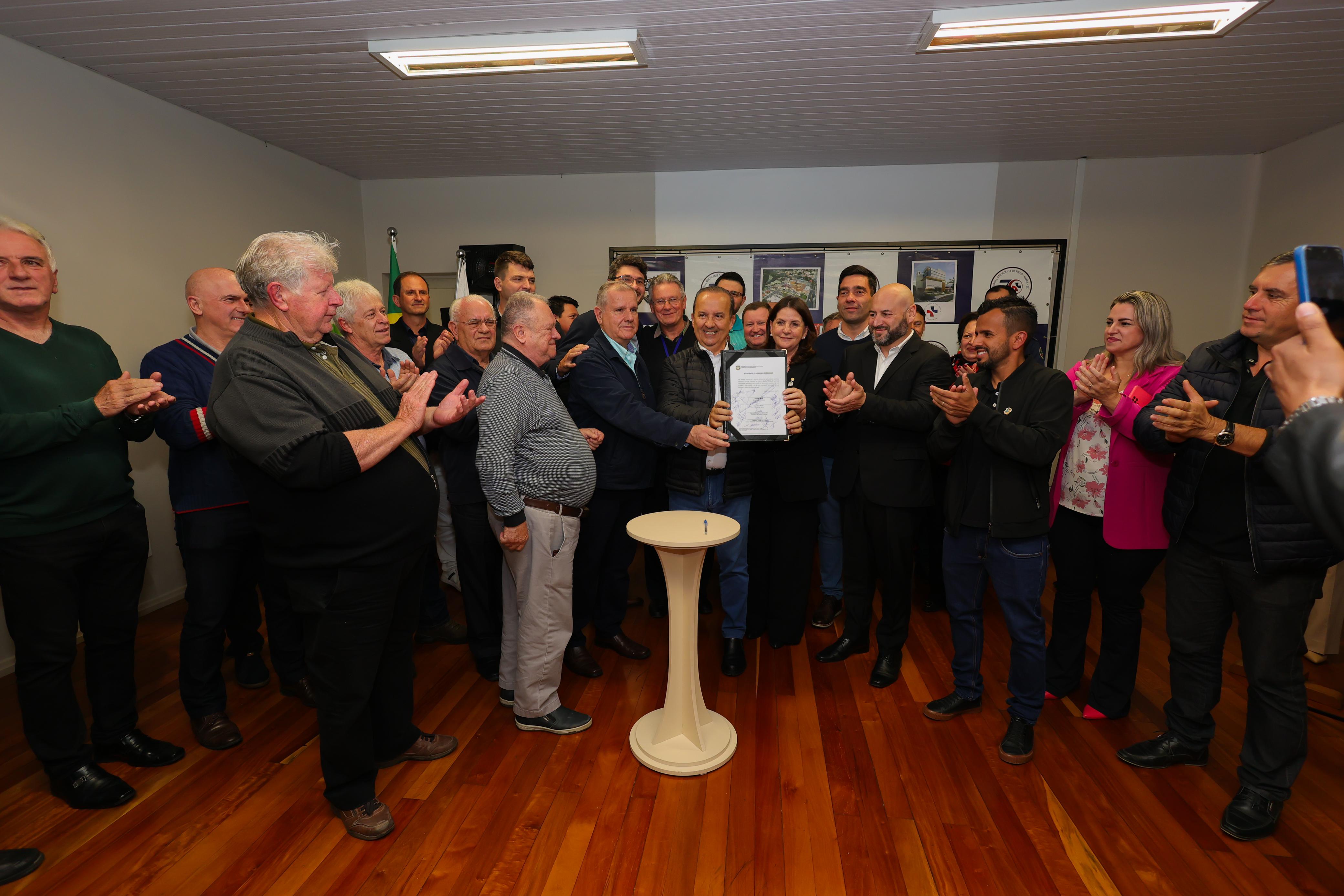 Grupo de pessoas reunidas em sala, aplaudindo enquanto autoridades seguram documento assinado de convênio do Hospital São Vicente de Paulo, em Mafra, com mesa de apoio e caneta à frente.