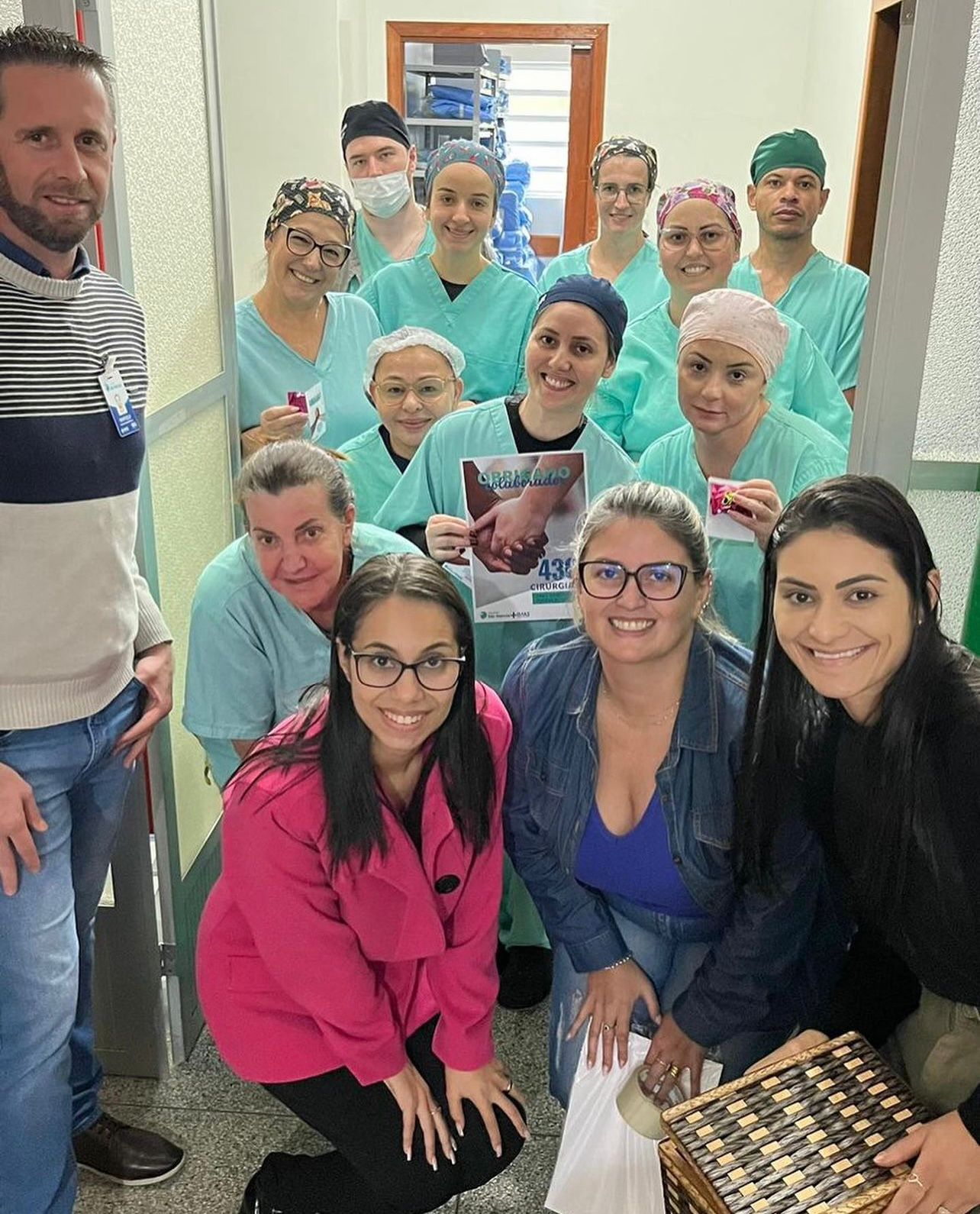 Grupo de profissionais da saúde posa sorridente em um corredor de hospital. A maioria veste roupas cirúrgicas verdes, toucas e máscaras (algumas abaixadas), enquanto três mulheres à frente estão em trajes casuais e seguram cestas e sacolas. Uma das profissionais segura um cartaz comemorativo indicando a realização de 438 cirurgias. Ambiente limpo e iluminado, com portas e armários hospitalares ao fundo.