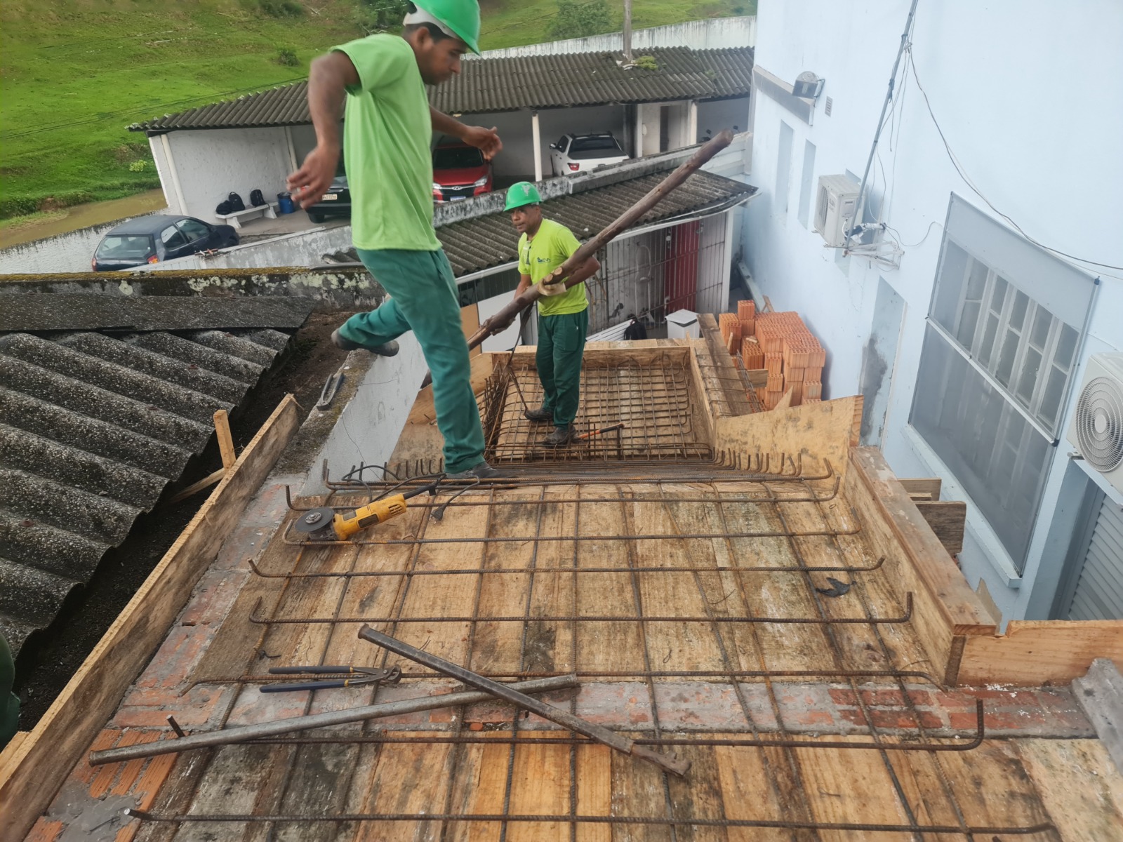 Dois trabalhadores com capacete verde e uniforme verde realizam serviços sobre uma laje em construção. Um deles caminha sobre a estrutura metálica, enquanto o outro segura uma barra de ferro. A imagem mostra o cuidado necessário em altura e o uso de equipamentos de proteção individual.
