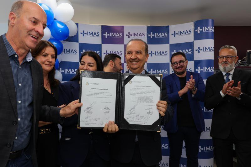 Grupo de pessoas sorriem durante cerimônia de assinatura de convênio, com destaque para homem ao centro exibindo documento oficial assinado. Fundo decorado com painel do IMAS e balões azuis e brancos.