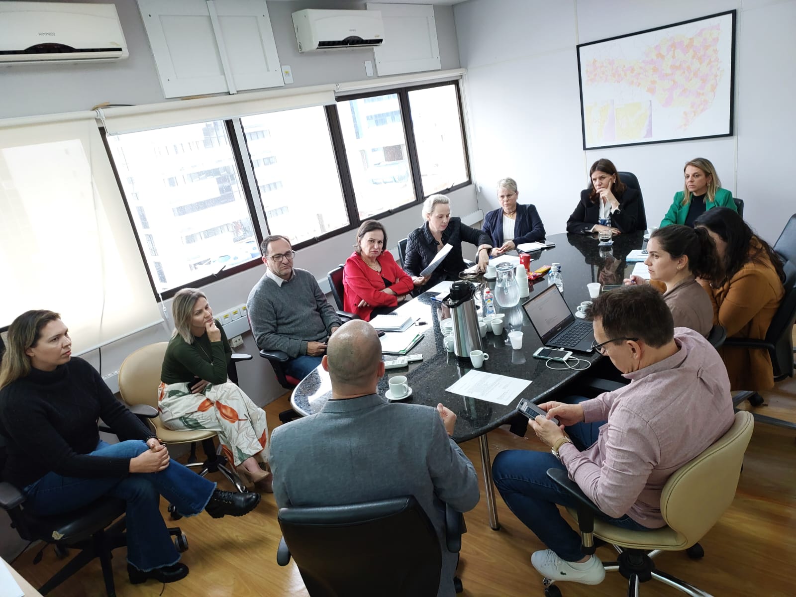 Grupo de profissionais de saúde reunidos em torno de uma mesa de reuniões em ambiente corporativo, discutindo temas relacionados a UTIs pediátricas. Há laptops, papéis e xícaras de café sobre a mesa.