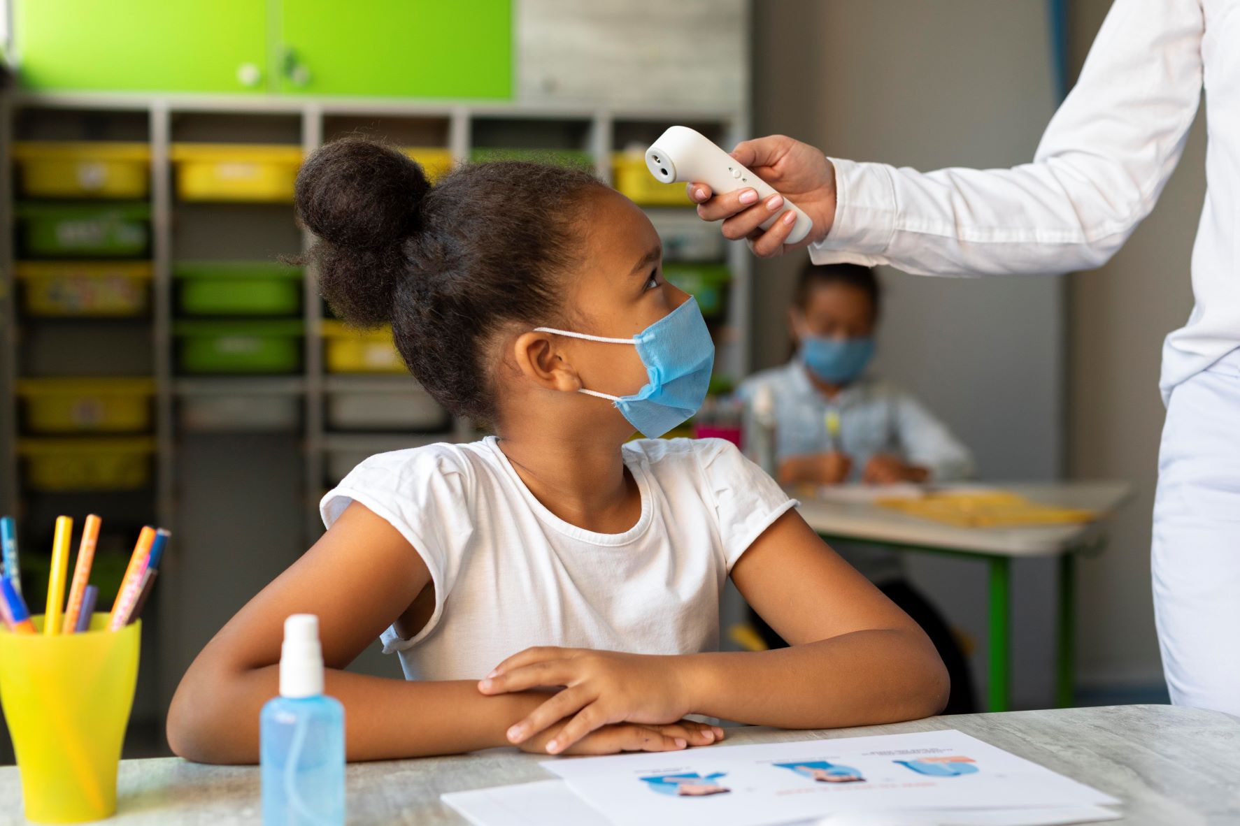 Criança usando máscara facial azul sentada em sala de aula, com as mãos cruzadas sobre a mesa. Uma pessoa, fora de foco, mede sua temperatura com um termômetro infravermelho. Na mesa há um frasco de álcool em gel, lápis coloridos e uma folha com ilustrações sobre cuidados com a saúde. Ao fundo, outra criança também de máscara está sentada, escrevendo. A cena remete a protocolos de saúde em ambientes escolares.