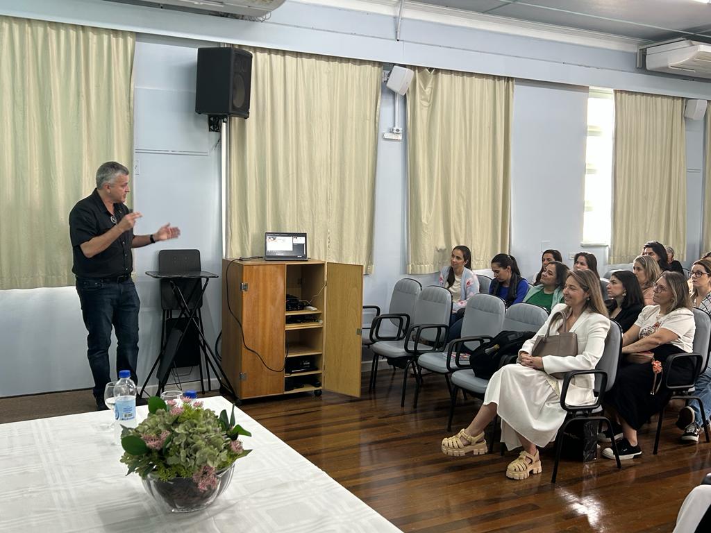 Palestrante apresenta conteúdo com apoio de notebook em uma sala de aula ou auditório. À direita, um grupo de mulheres atentas assiste à apresentação. No primeiro plano, há uma mesa com toalha branca e um arranjo floral. As cortinas estão fechadas e a luz natural entra pelas janelas.
