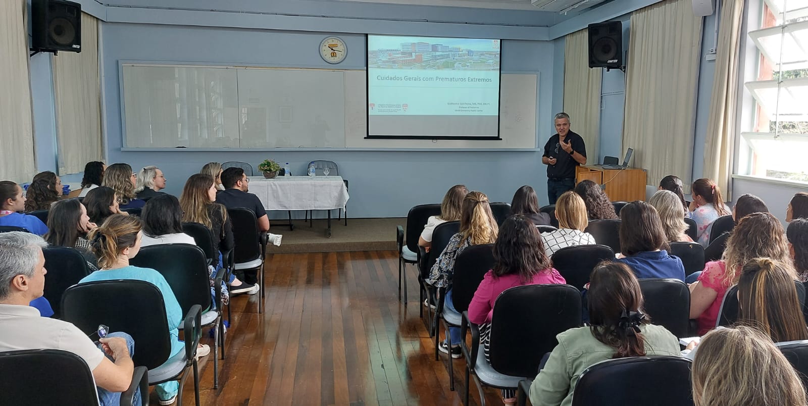 Palestra ministrada por profissional da saúde em auditório com diversas pessoas sentadas assistindo. Na tela de projeção, lê-se o título “Cuidados Gerais com Prematuros Extremos” e o nome do palestrante: Guilherme Barros, MD, PhD, MPH. O ambiente é claro, com cortinas claras e janelas grandes à direita, e um relógio acima do quadro branco.