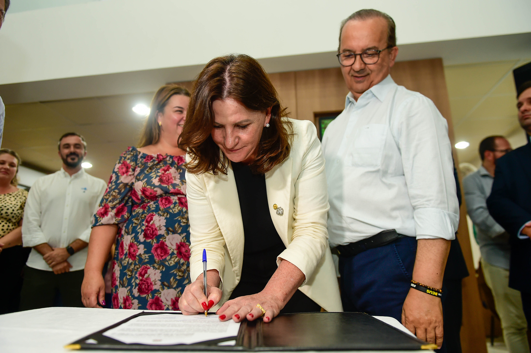 Mulher de blazer claro assina um documento sobre uma mesa branca, segurando uma caneta azul. Ao fundo, várias pessoas observam o momento, incluindo um homem de óculos ao lado direito e uma mulher com vestido floral sorrindo. A cena ocorre em ambiente fechado, com iluminação artificial.