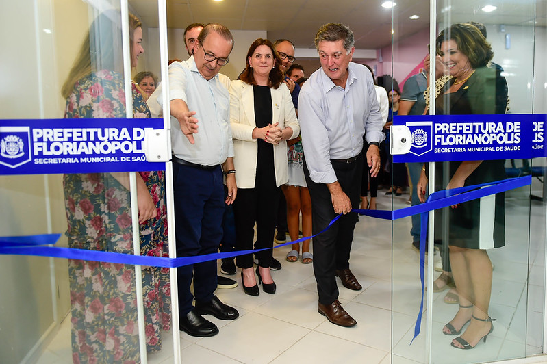 Grupo de pessoas participa da cerimônia de inauguração de uma unidade da Secretaria Municipal de Saúde da Prefeitura de Florianópolis. Um homem, à frente, corta uma fita azul simbolizando a abertura oficial. Ao seu lado, uma mulher de blazer claro e outros participantes observam sorridentes. A cena ocorre em ambiente interno, com piso branco e portas de vidro com adesivos institucionais.