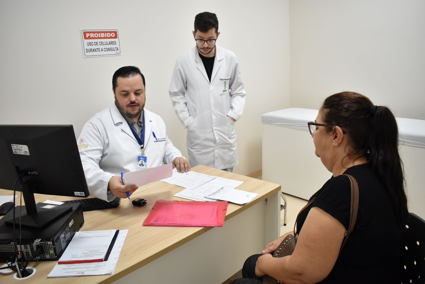Médico e estagiário, ambos de jaleco branco, atendem uma paciente em consultório. O médico segura um papel e conversa com a paciente, enquanto o outro profissional observa. Na parede, há uma placa com a mensagem "Proibido uso de celulares durante a consulta".
