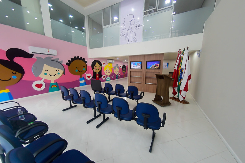 Sala de acolhimento infantil com decoração colorida e temática voltada para o público infantil. O espaço possui cadeiras organizadas em fileiras, um púlpito de madeira ao lado de bandeiras institucionais, e paredes ilustradas com desenhos de crianças sorridentes. No fundo, há uma estante com telas digitais e, acima, uma imagem delicada de uma mãe com bebê. O ambiente é iluminado e moderno, com piso branco e vidro no mezanino superior.