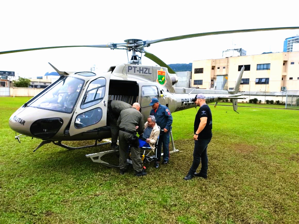 Helicóptero da Polícia Militar de Santa Catarina (PT-HZL) estacionado em área gramada, com equipe de resgate auxiliando no embarque de um paciente em cadeira de rodas. A operação envolve profissionais do SAMU e da PM, demonstrando integração para transporte aeromédico.
