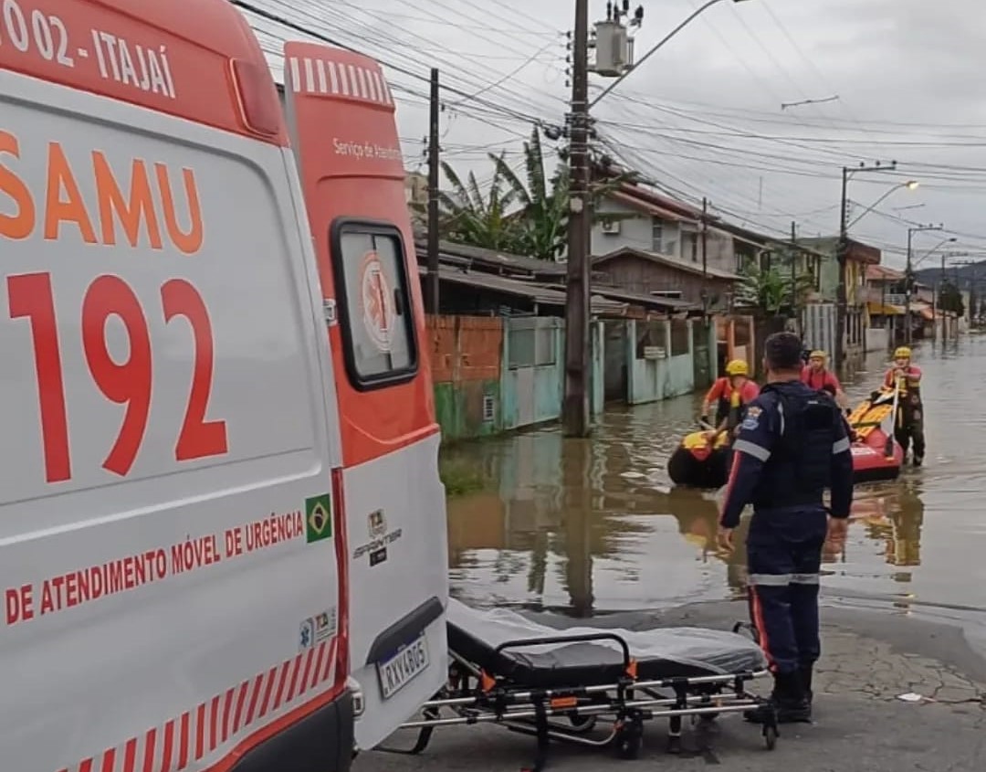Ambulância do SAMU estacionada próxima a rua alagada em Itajaí, com maca no chão e equipe de resgate transportando vítima em bote inflável.